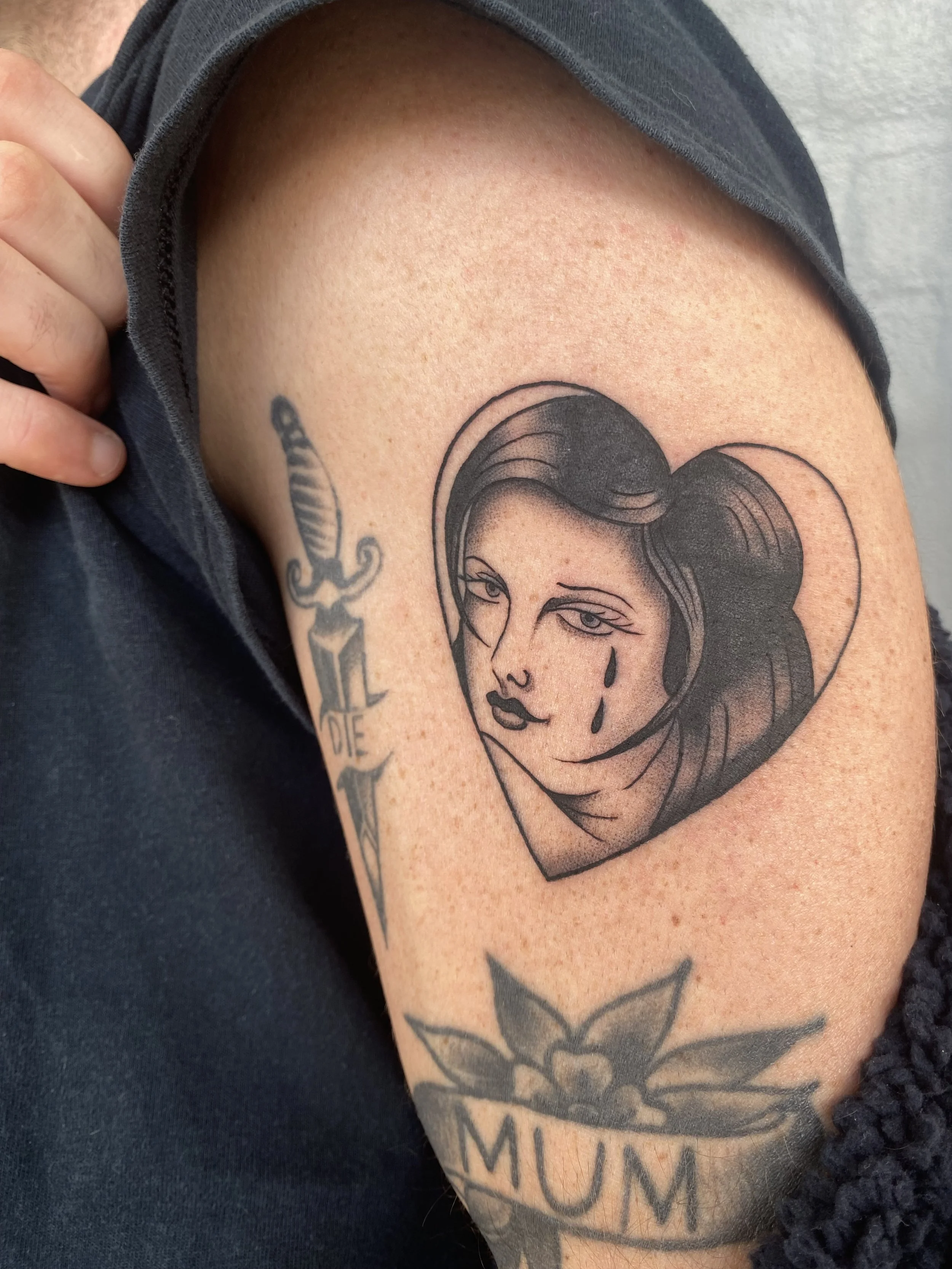 Tattoo of a woman's face with a tear on her cheek inside a heart, on upper arm of person wearing black clothing.