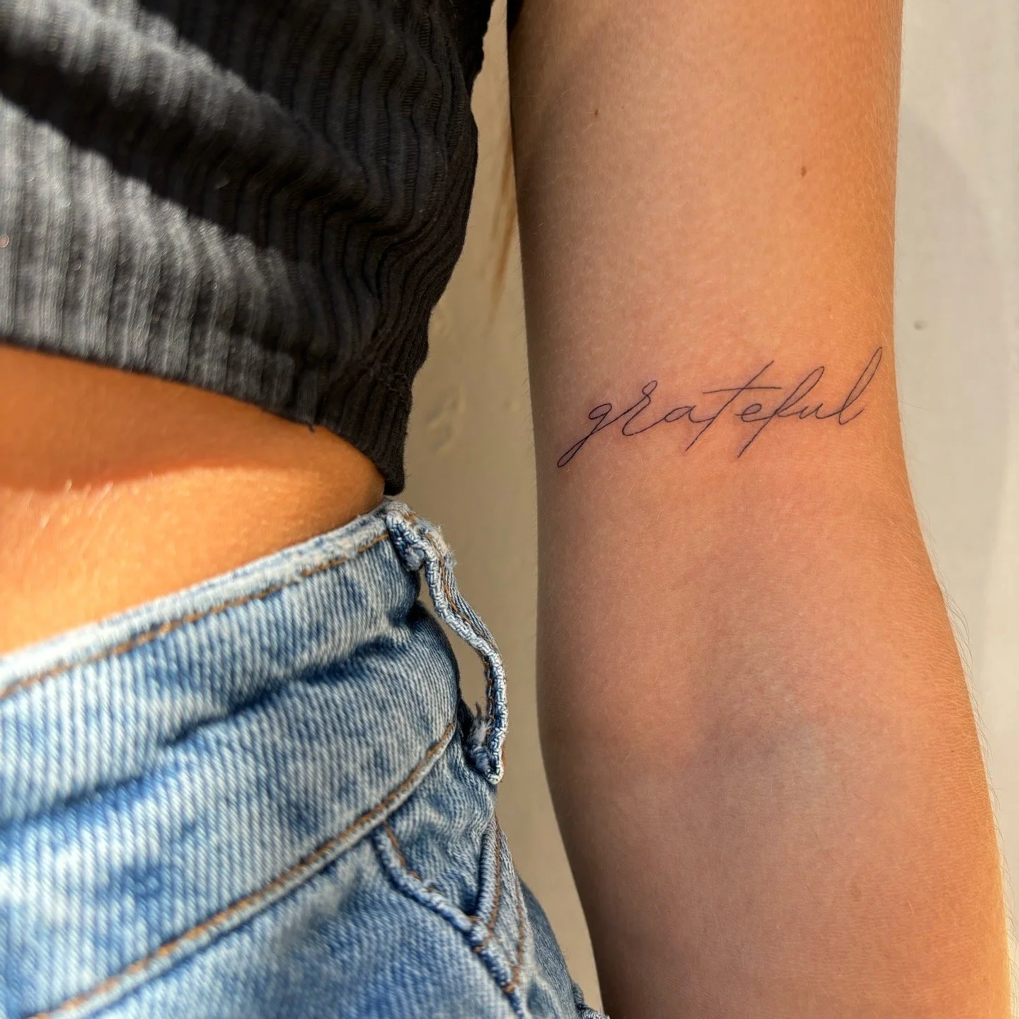 A close-up of a person's arm with a tattoo that says "grateful" in cursive script. The person is wearing a dark, ribbed top and light blue jeans.
