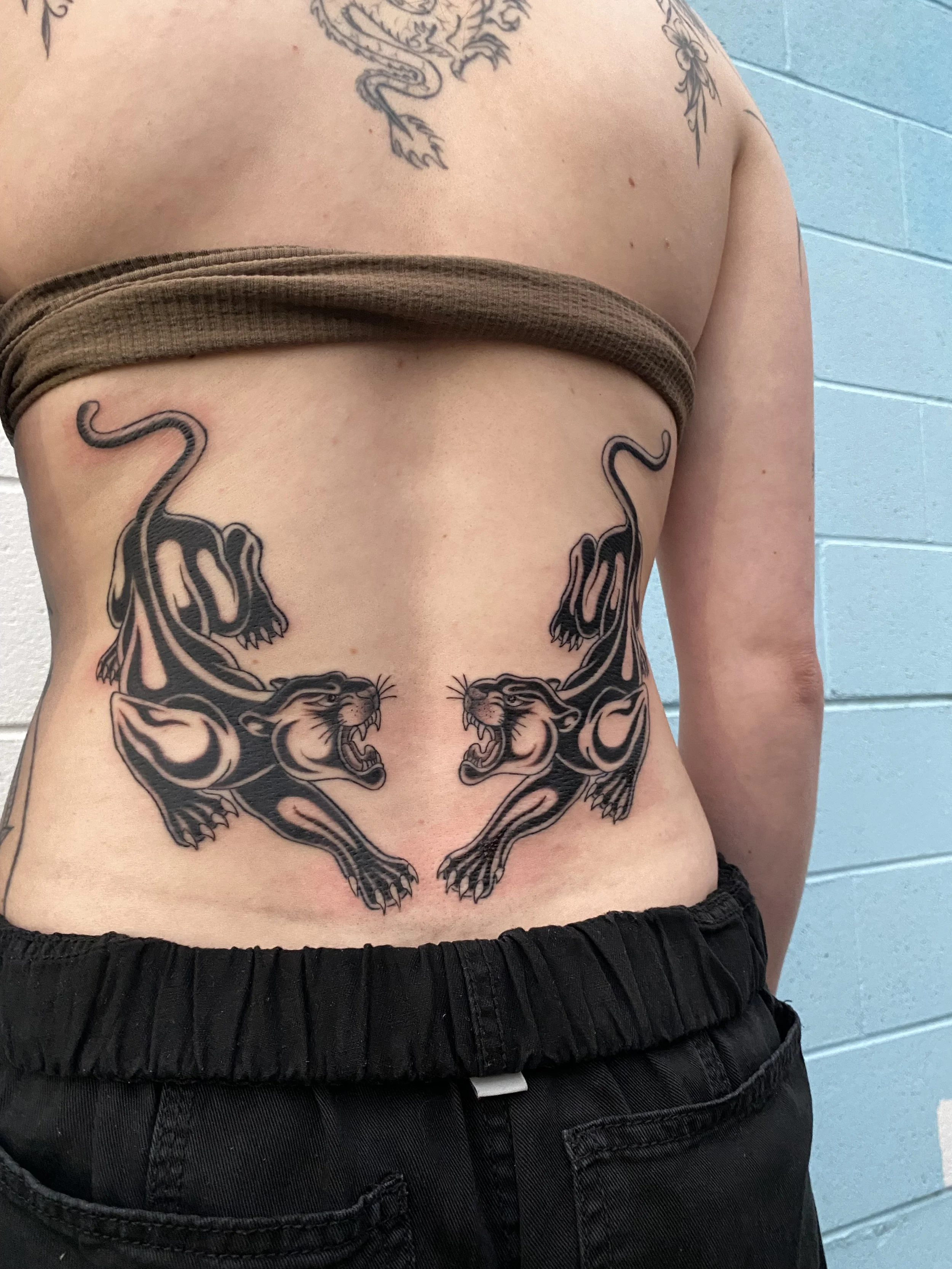 Tattoo of two black panthers with open mouths, facing each other, on a person's lower abdomen and sides.