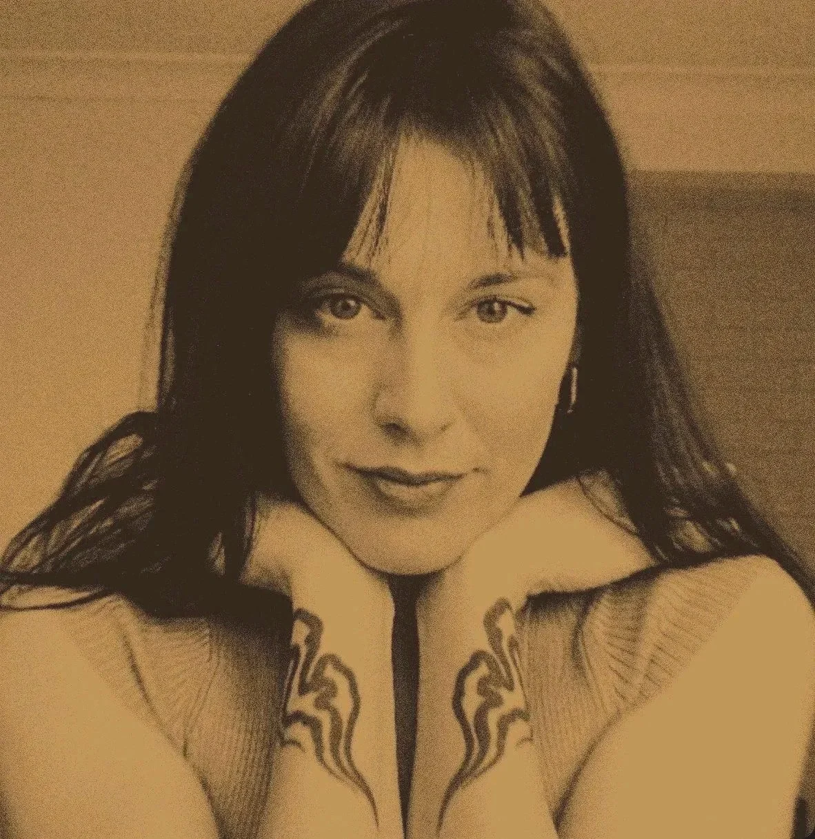 Black and white portrait of a woman with straight dark hair and bangs, resting her chin on her hands, looking at the camera with a slight smile, showing tattoos on her forearms.
