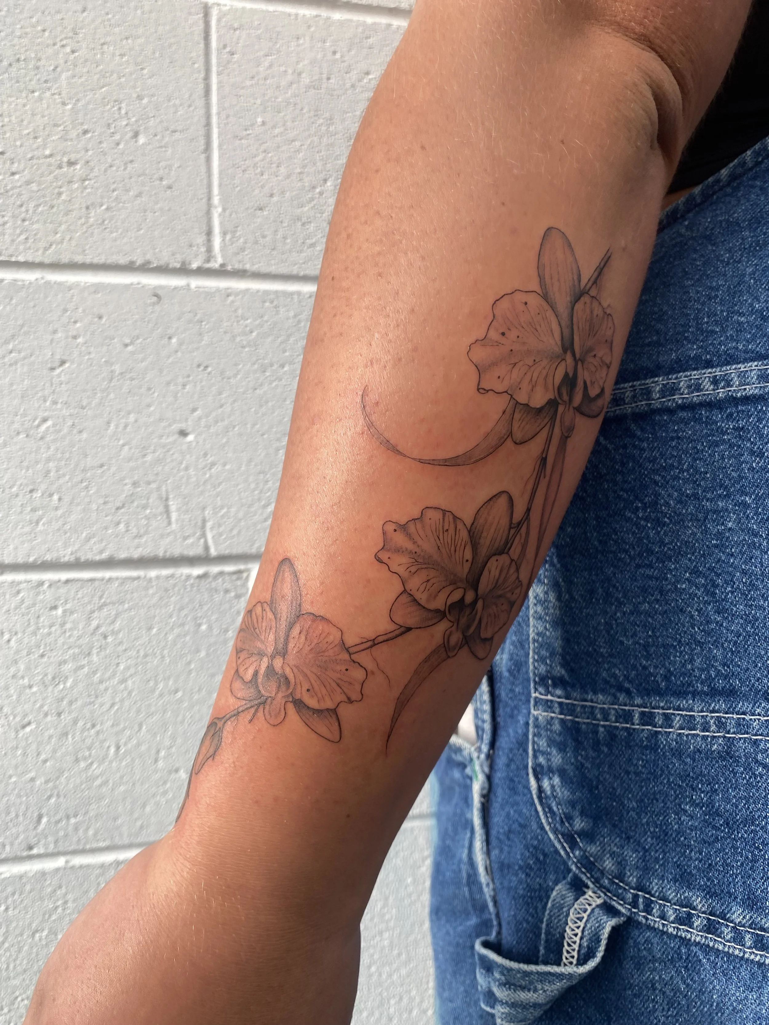 Black ink line tattoo of two orchids on inner forearm near the elbow, next to a person wearing blue jeans.