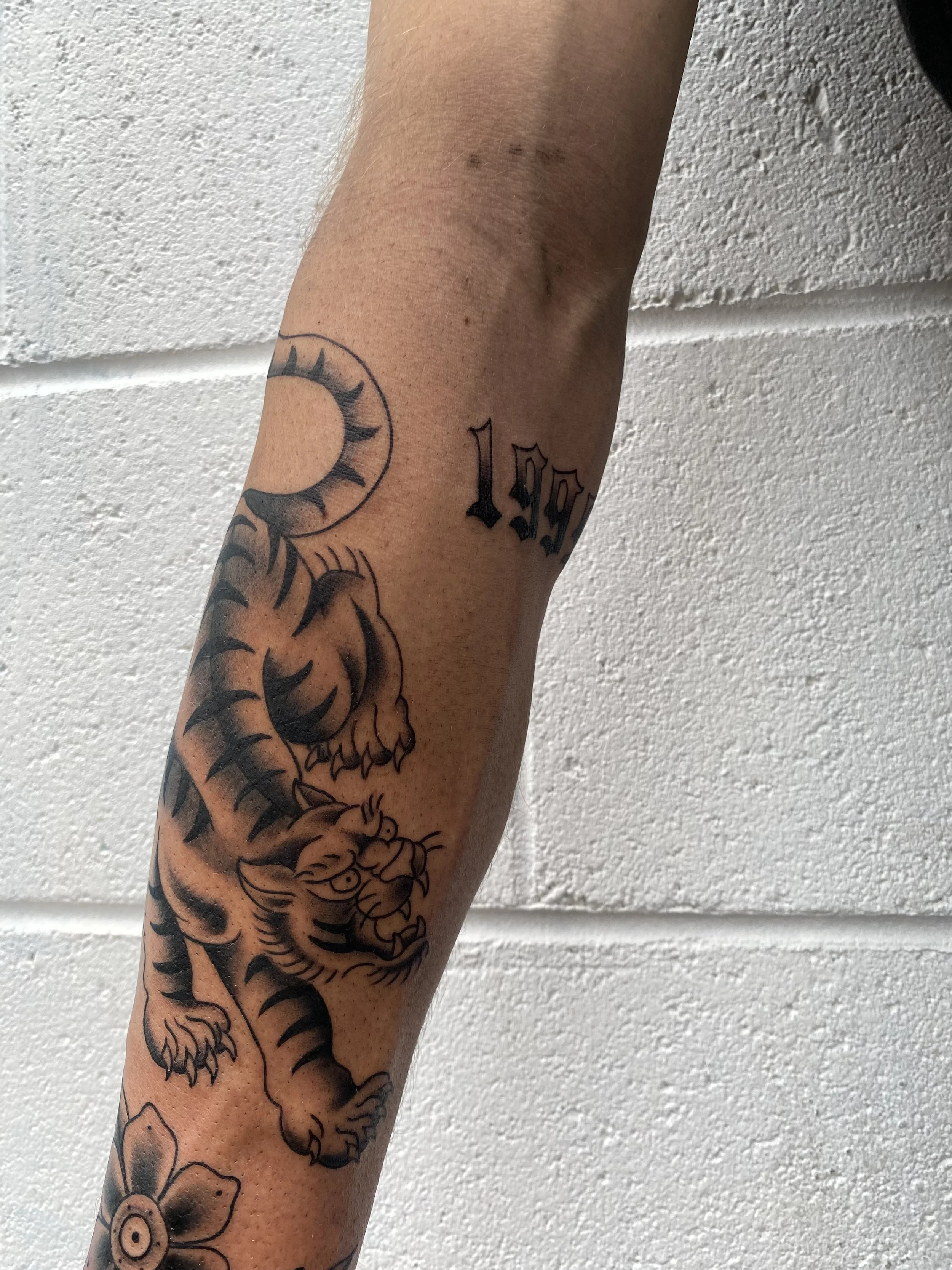 Tattoo of a tiger and a crescent moon with the year 1999 on a person's arm against a white brick wall background.