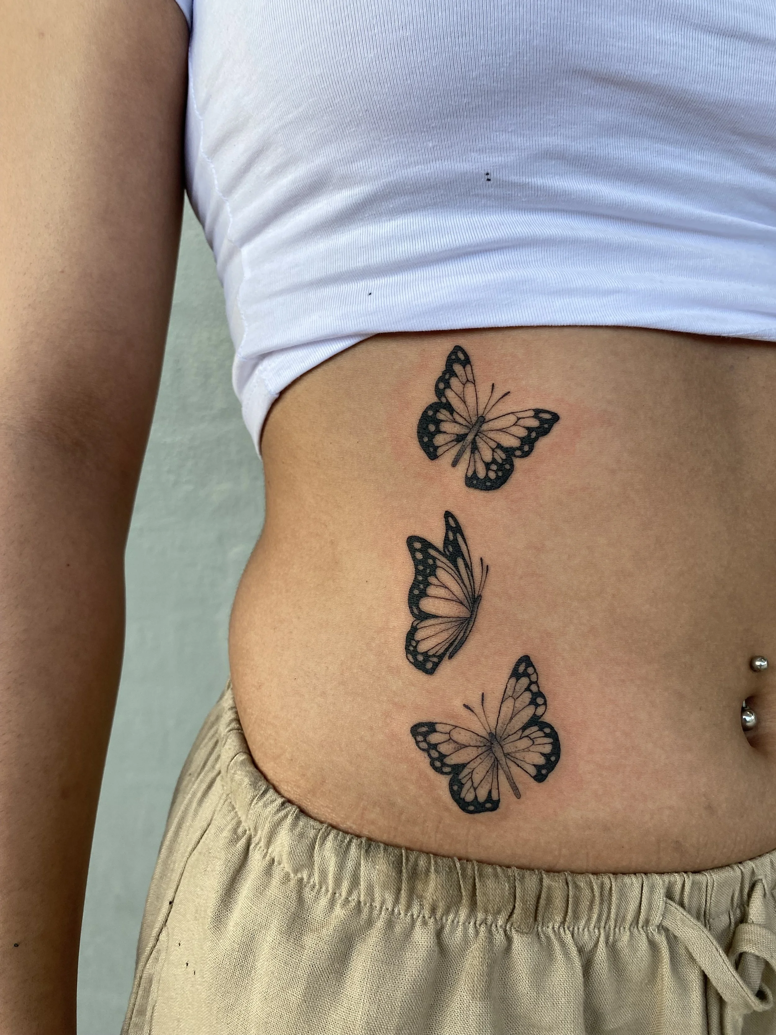 Tattoo of three butterflies on the person's side, above beige shorts and below a white crop top.