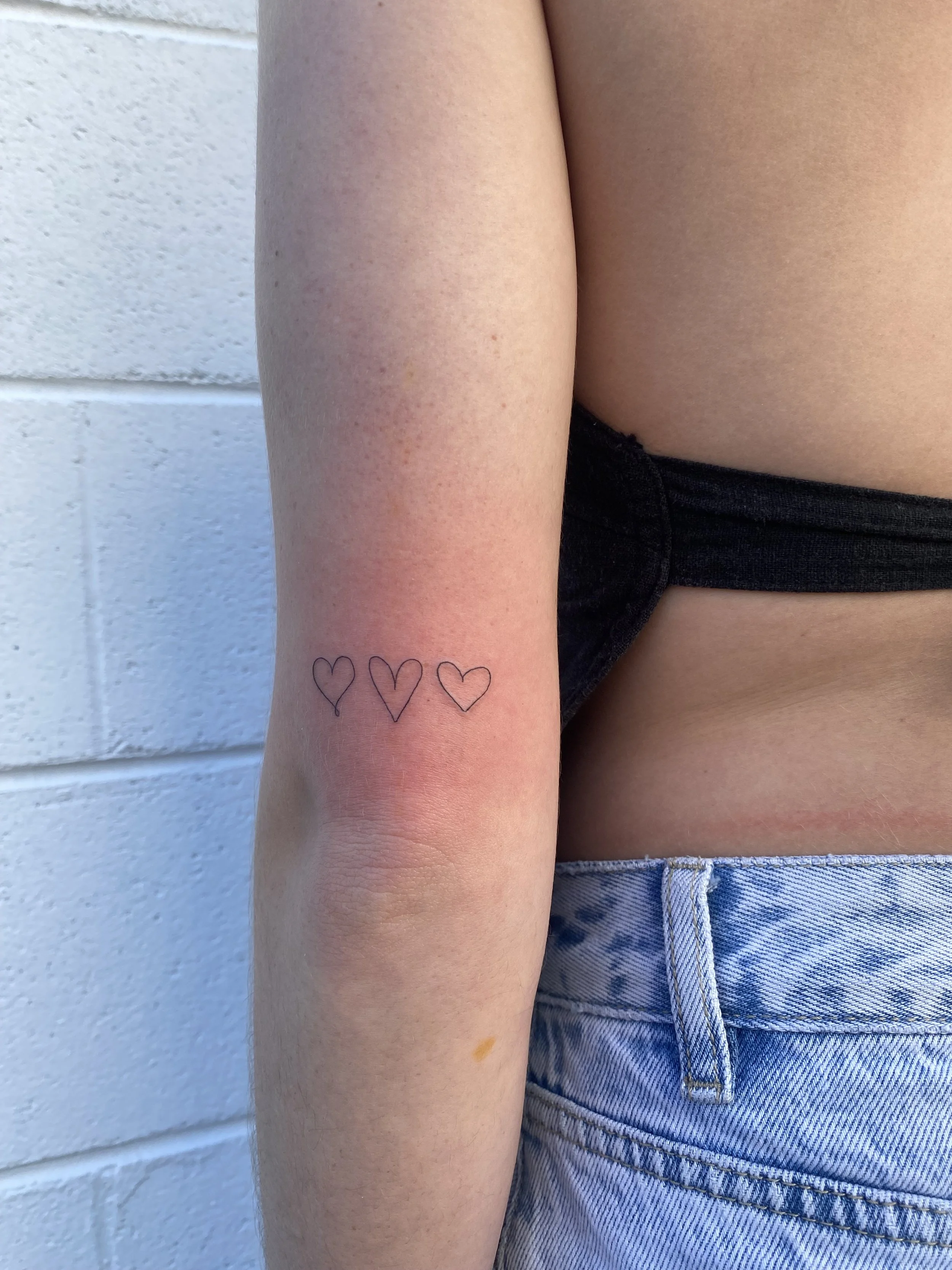 Close-up of a person's arm with a small tattoo of three heart outlines on the inner forearm, wearing a black top and light blue jeans against a white brick wall.