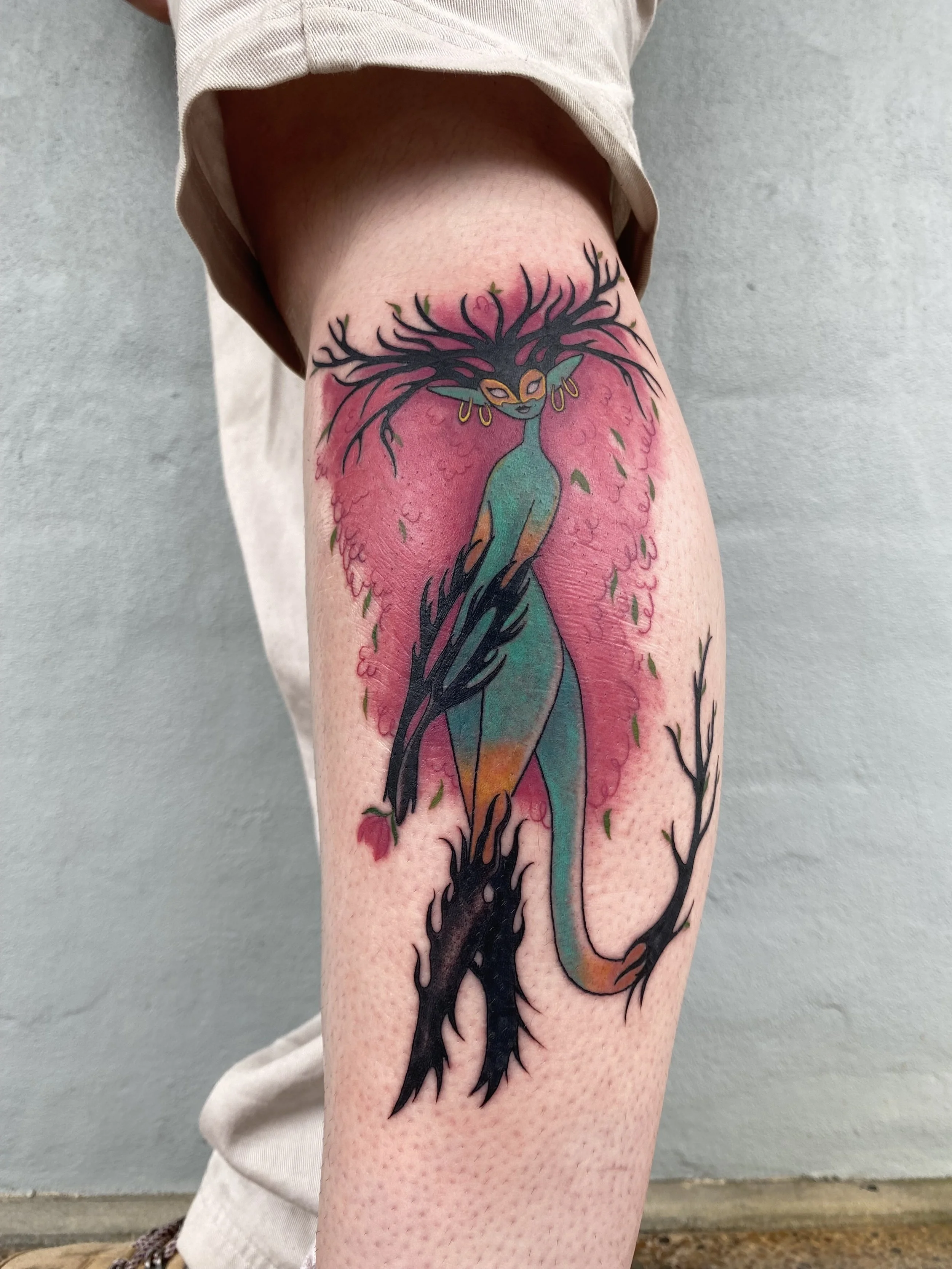 Colorful tattoo of a woman with flowing hair resembling tree branches, wearing earrings, with a pink and green background, on a person's thigh.