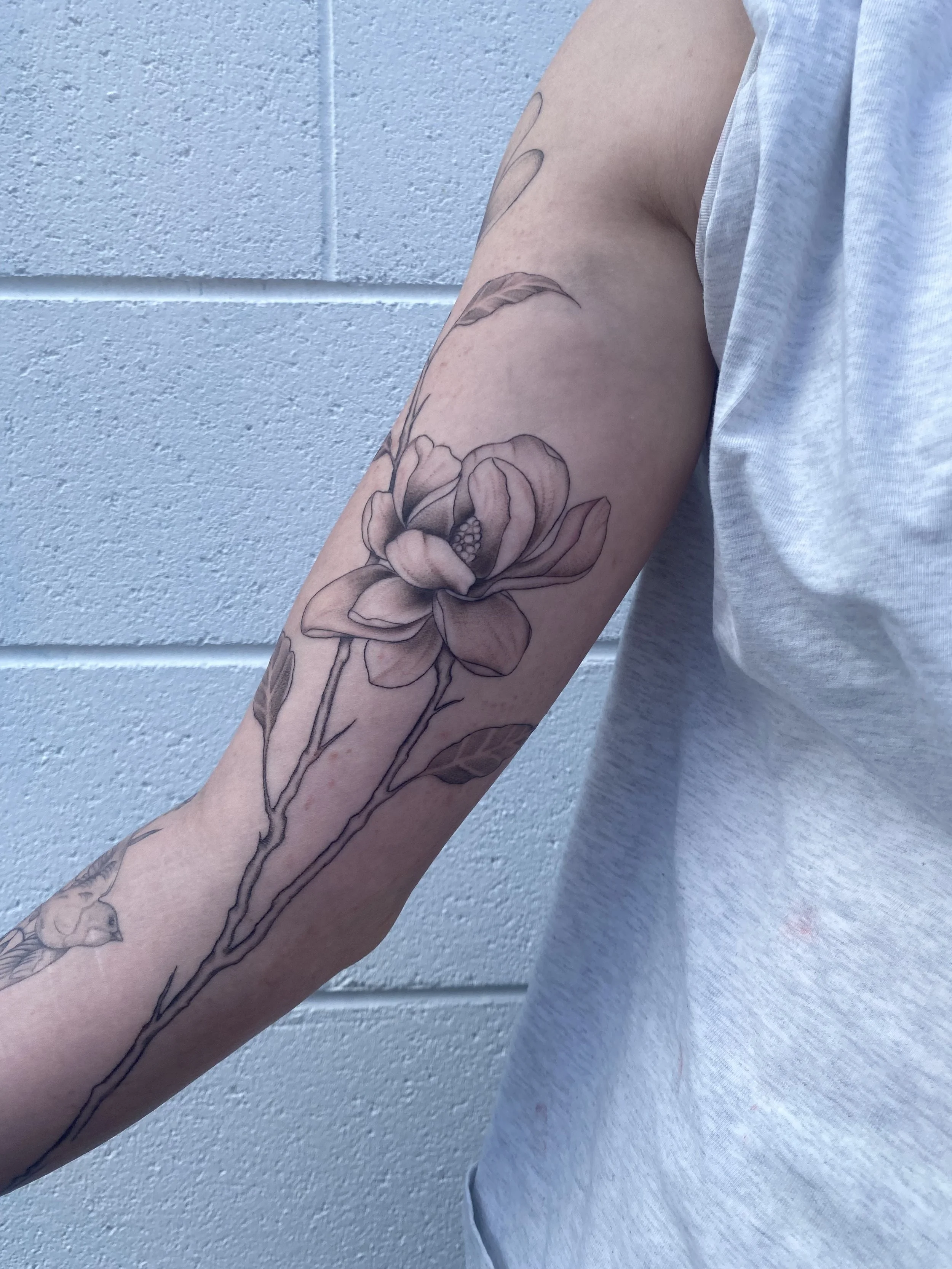 Tattoo of a large flower with leaves on a person's upper arm, in front of a light-colored brick wall.