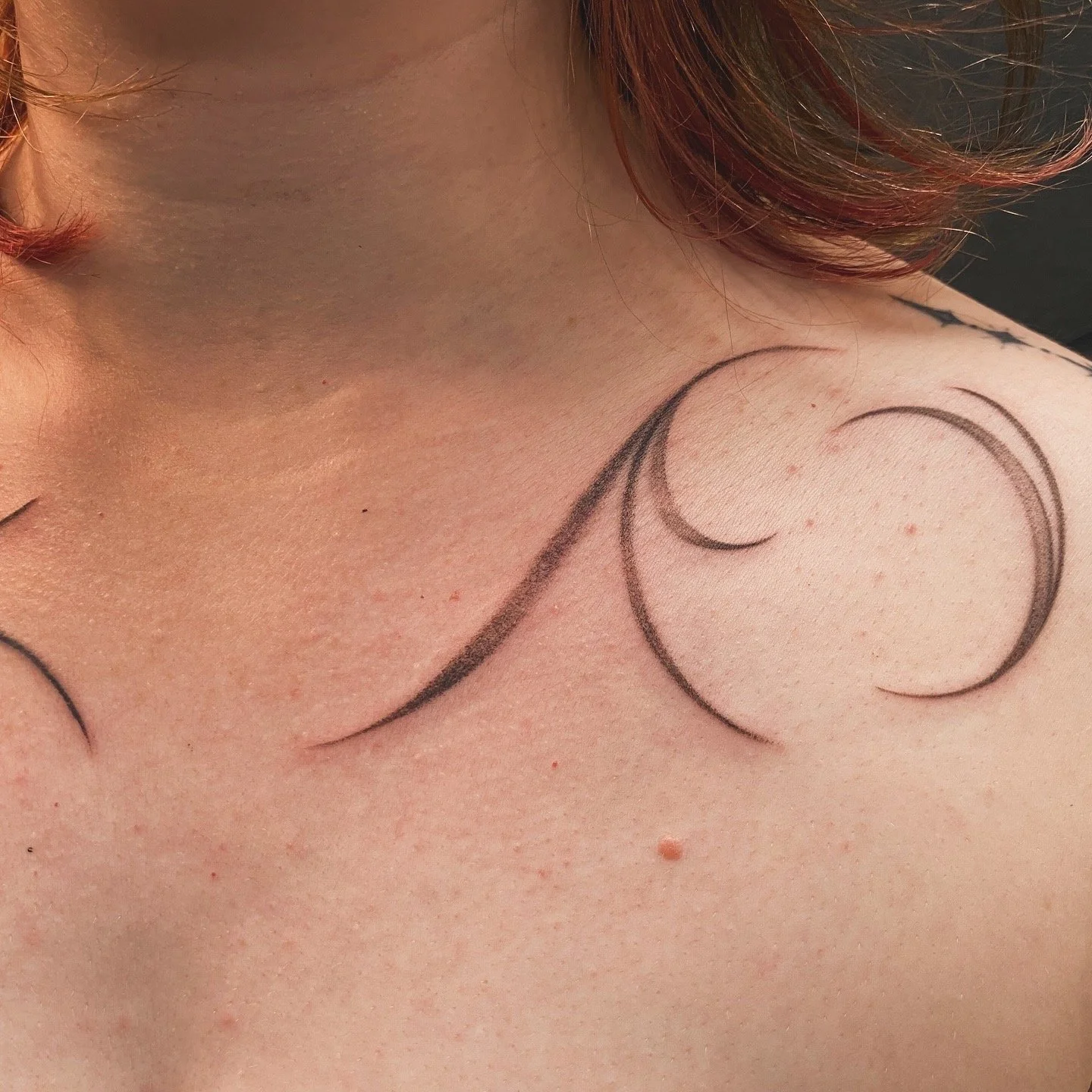 A close-up of a shoulder and collarbone with a black ink tattoo of swirling lines and crescent moons.