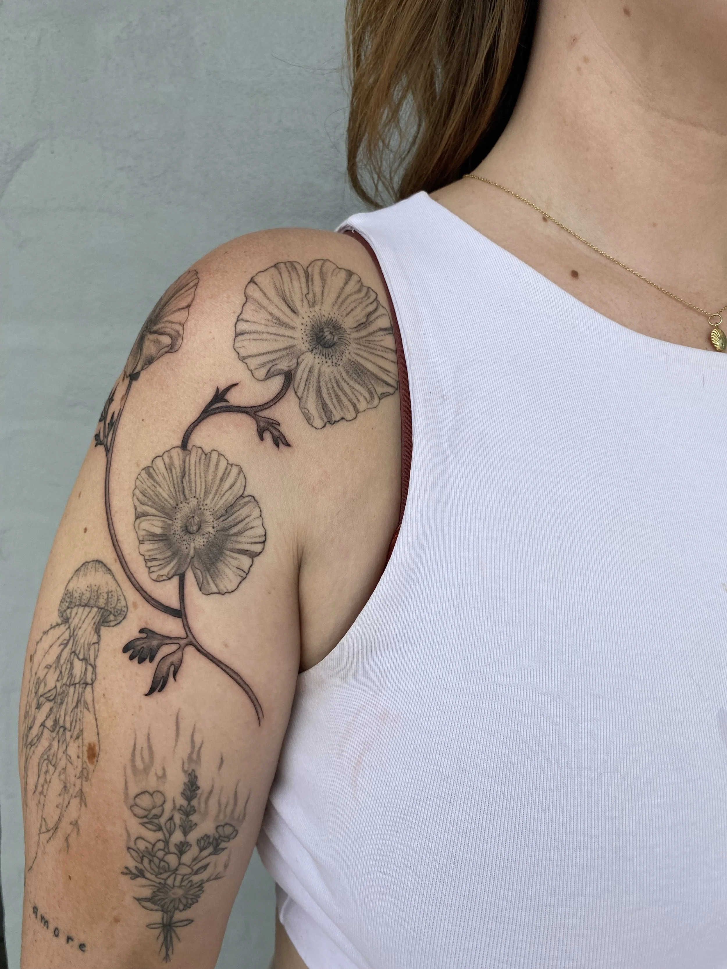 A person's upper arm and shoulder with a tattoo of a flowering plant and a jellyfish, wearing a white sleeveless top.
