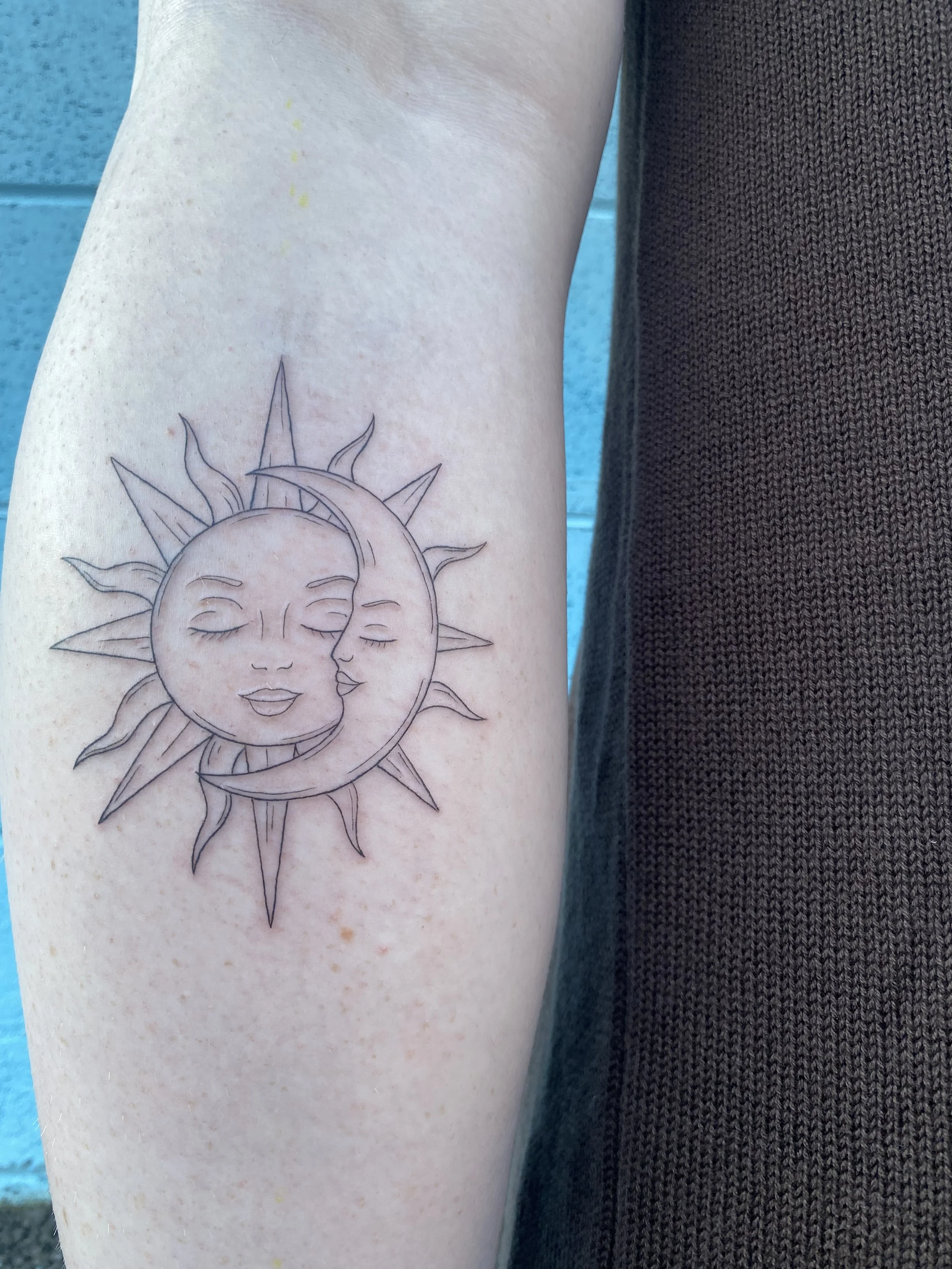Tattoo of a smiling sun with a face and a smiling moon with a face, intertwined and surrounded by rays of sunlight, on a person's upper arm.