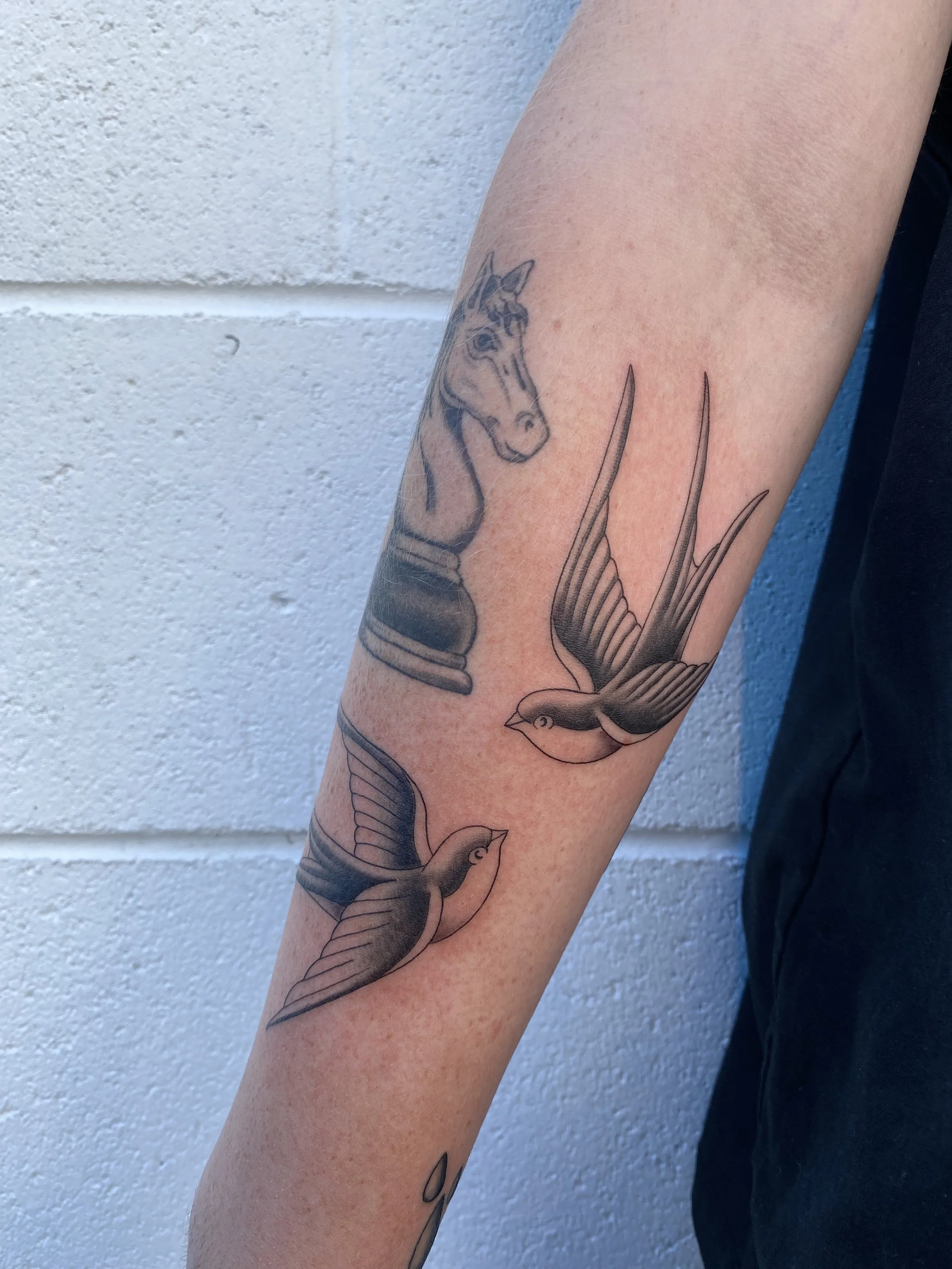 Tattoo of a chess knight piece and three flying birds on a person's forearm, with a plain white wall in the background.
