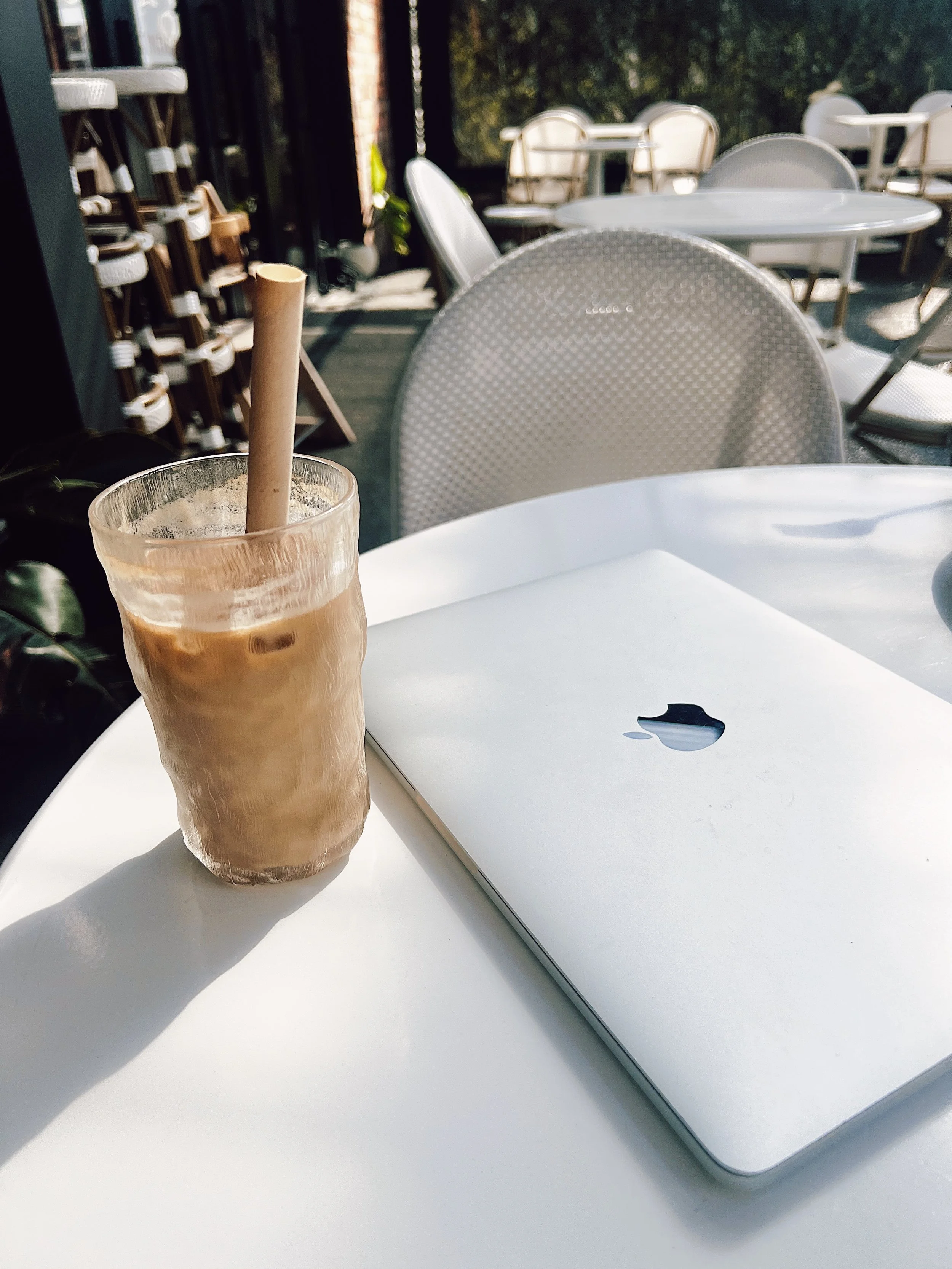 A glass of iced coffee with a paper straw and a closed silver Apple MacBook on a white outdoor table.