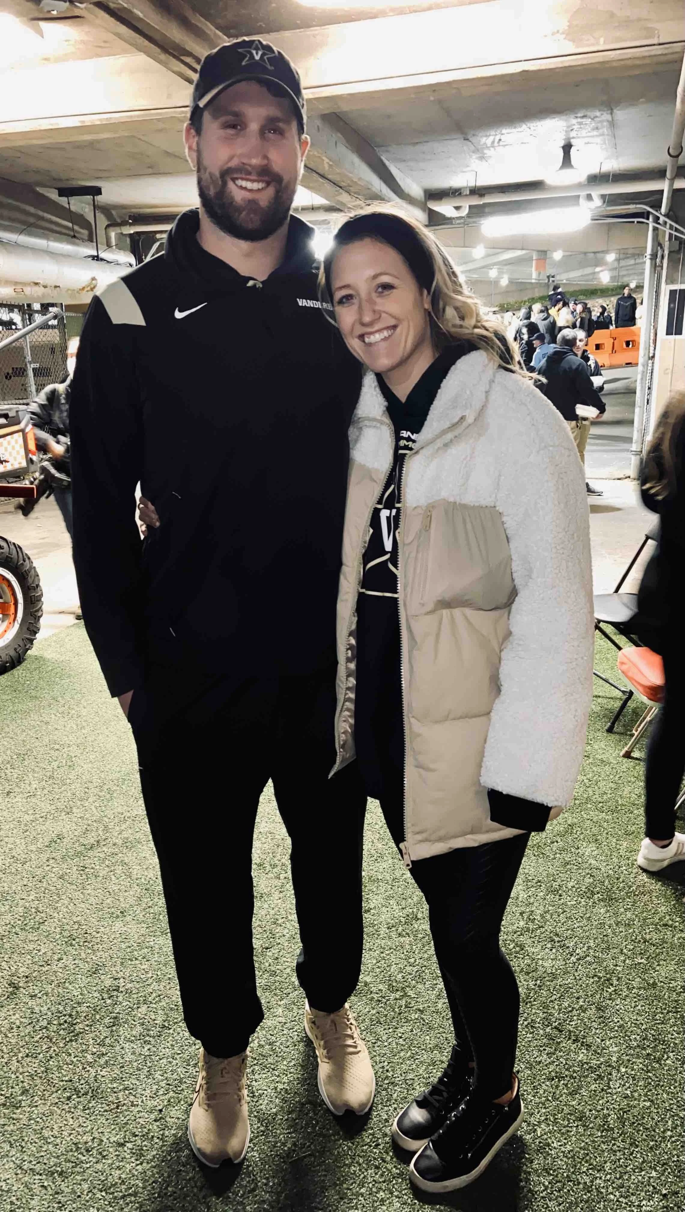 A man and woman standing together indoors, smiling at the camera, with a crowd in the background.