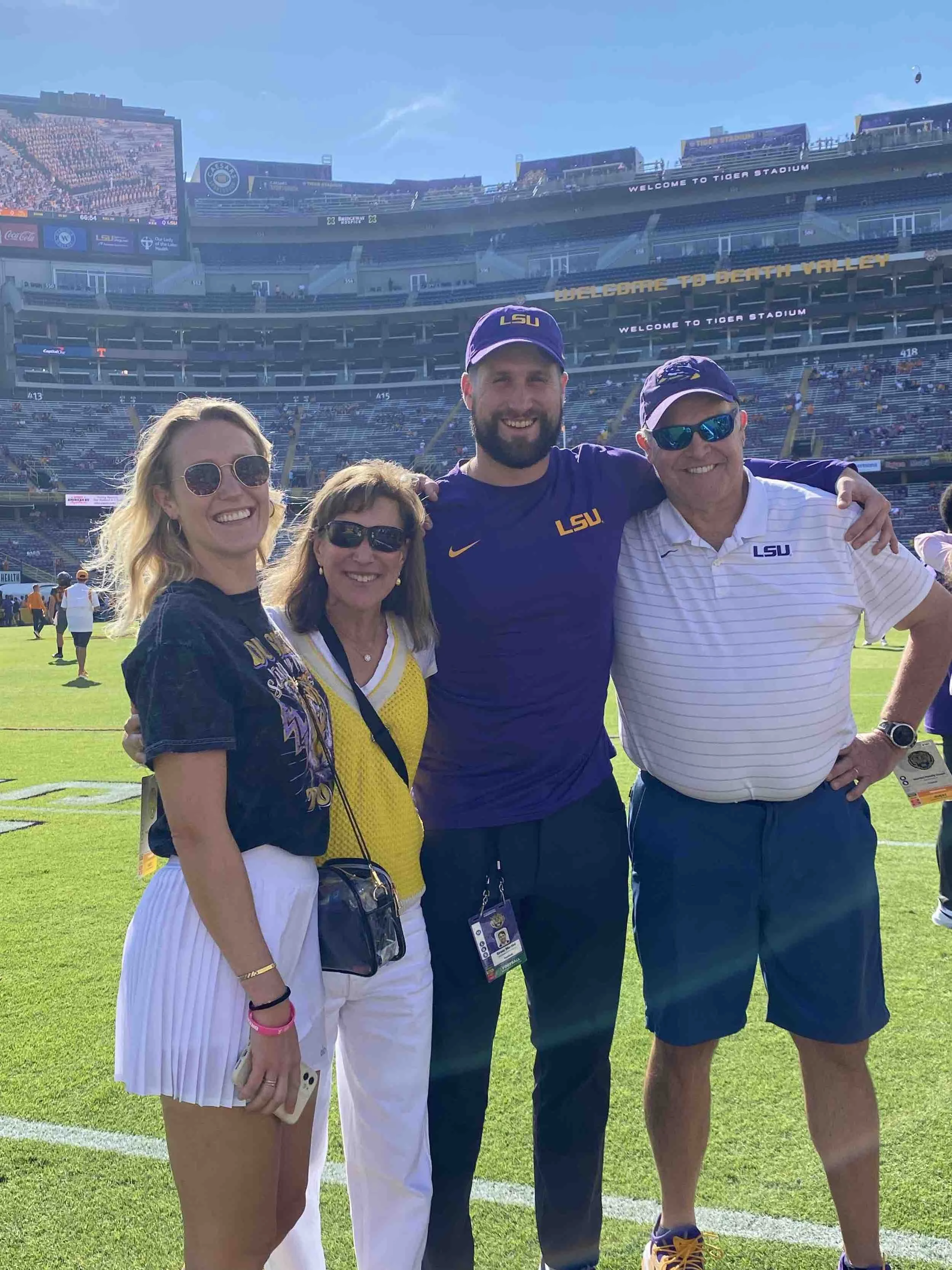 Group of four people on a football field in a stadium, smiling and posing for a photo. They are wearing LSU apparel and sunglasses, with clear skies overhead.
