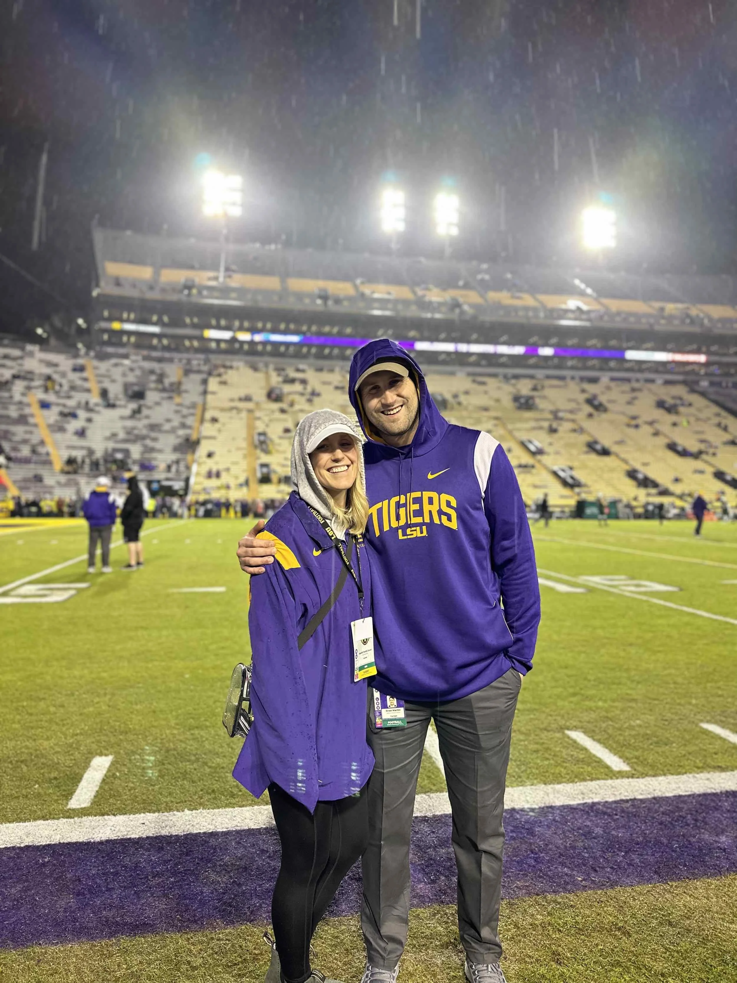 A smiling couple on a rainy night at a football stadium, wearing LSU Tigers apparel, standing on the field with stadium seats and floodlights in the background.