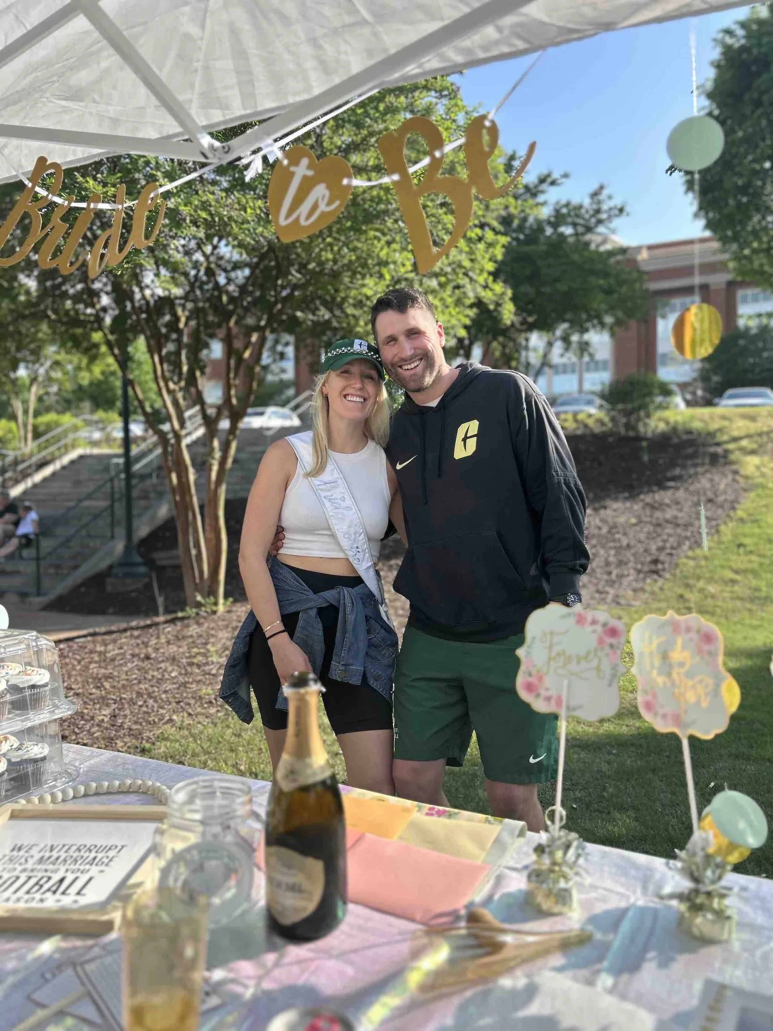 A smiling couple at a wedding reception outdoors under a canopy, with a 'Bride to Be' banner hanging above them and decorated tables with party favors in the foreground.