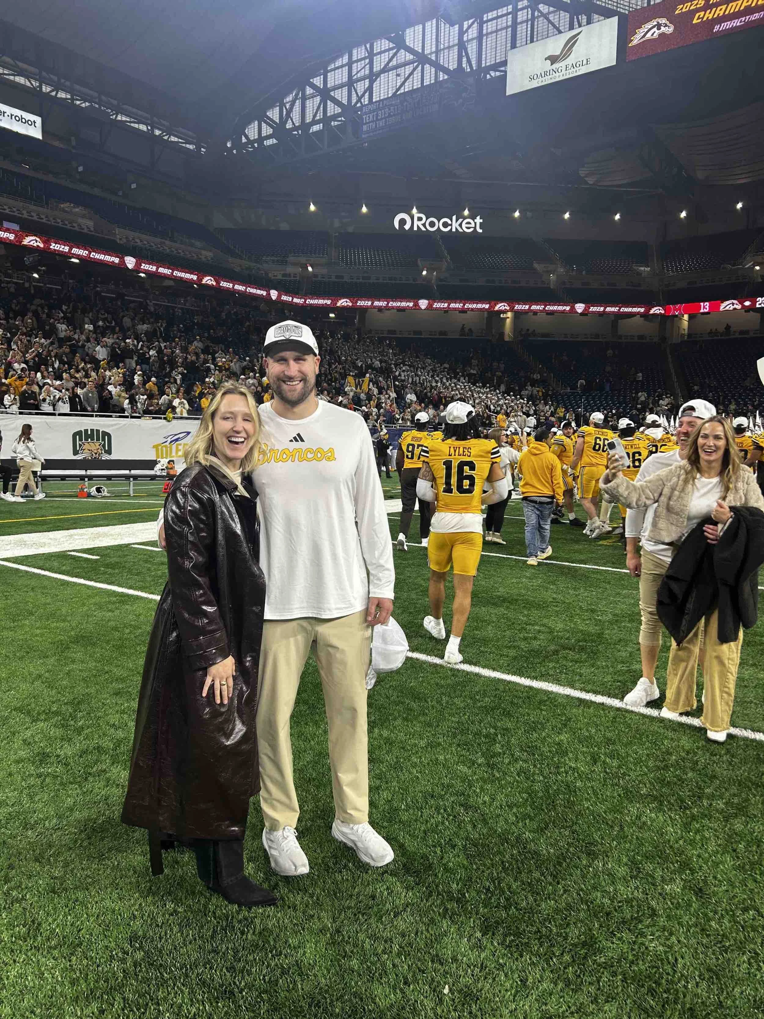 Two people smiling and standing on a football field, with a team in yellow uniforms and a crowd in the background. The woman wears a black trench coat, and the man wears a white Broncos shirt and beige pants.