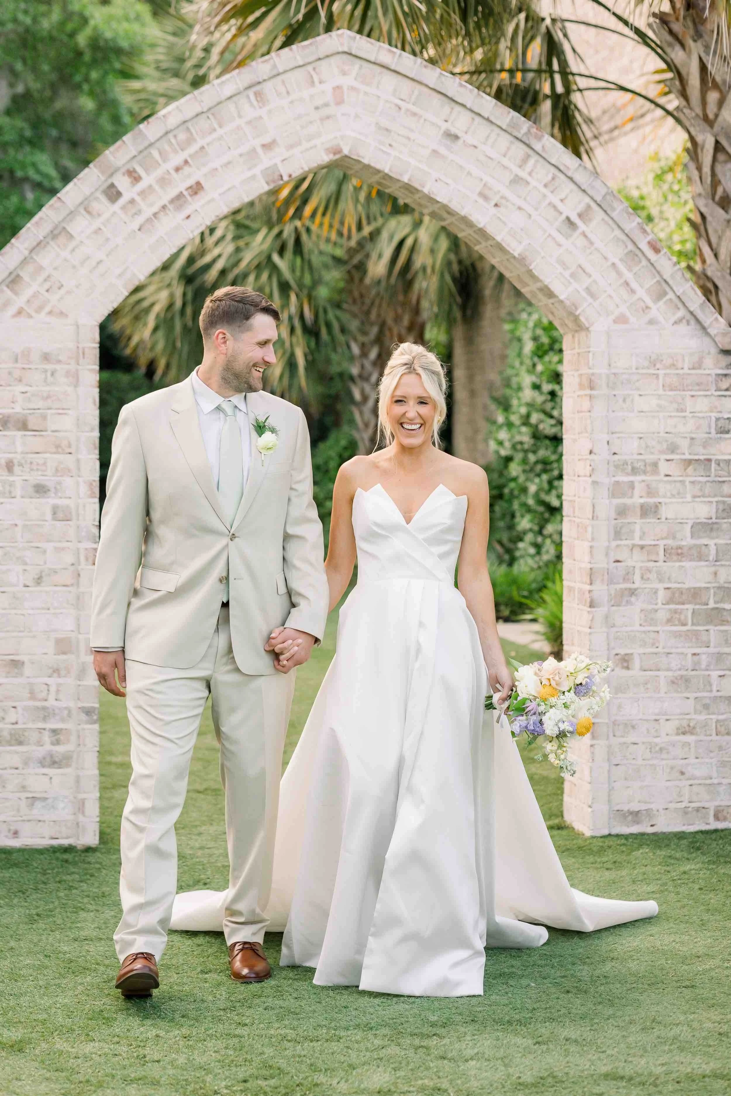 A bride and groom walking hand in hand under a white brick archway outdoors, smiling happily. The bride is in a white strapless wedding dress and holding a bouquet, while the groom is in a light-colored suit with a boutonniere. Green trees and plants surround them.