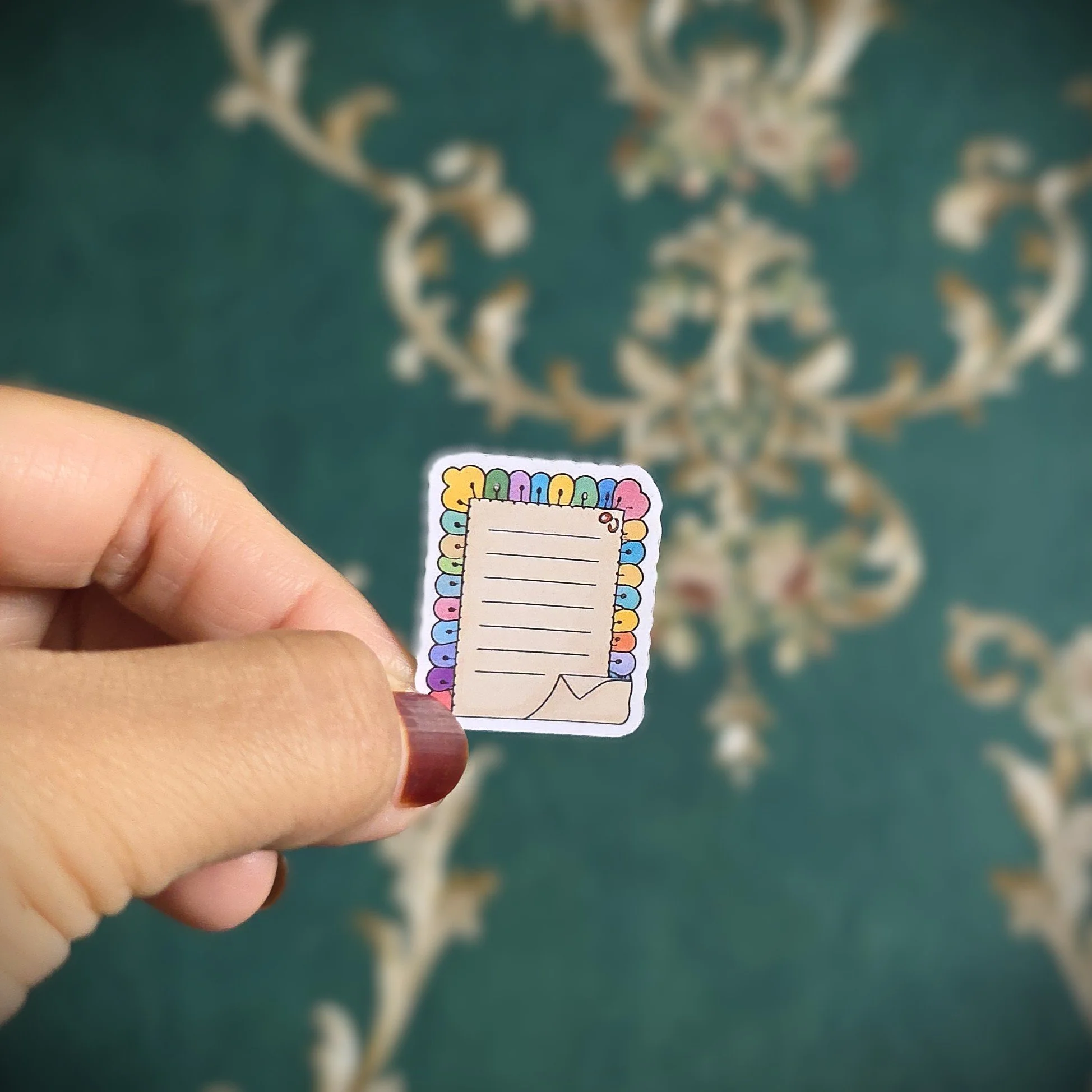 A person holding a small colorful notepad sticker with lined space for writing, against a green patterned carpet.