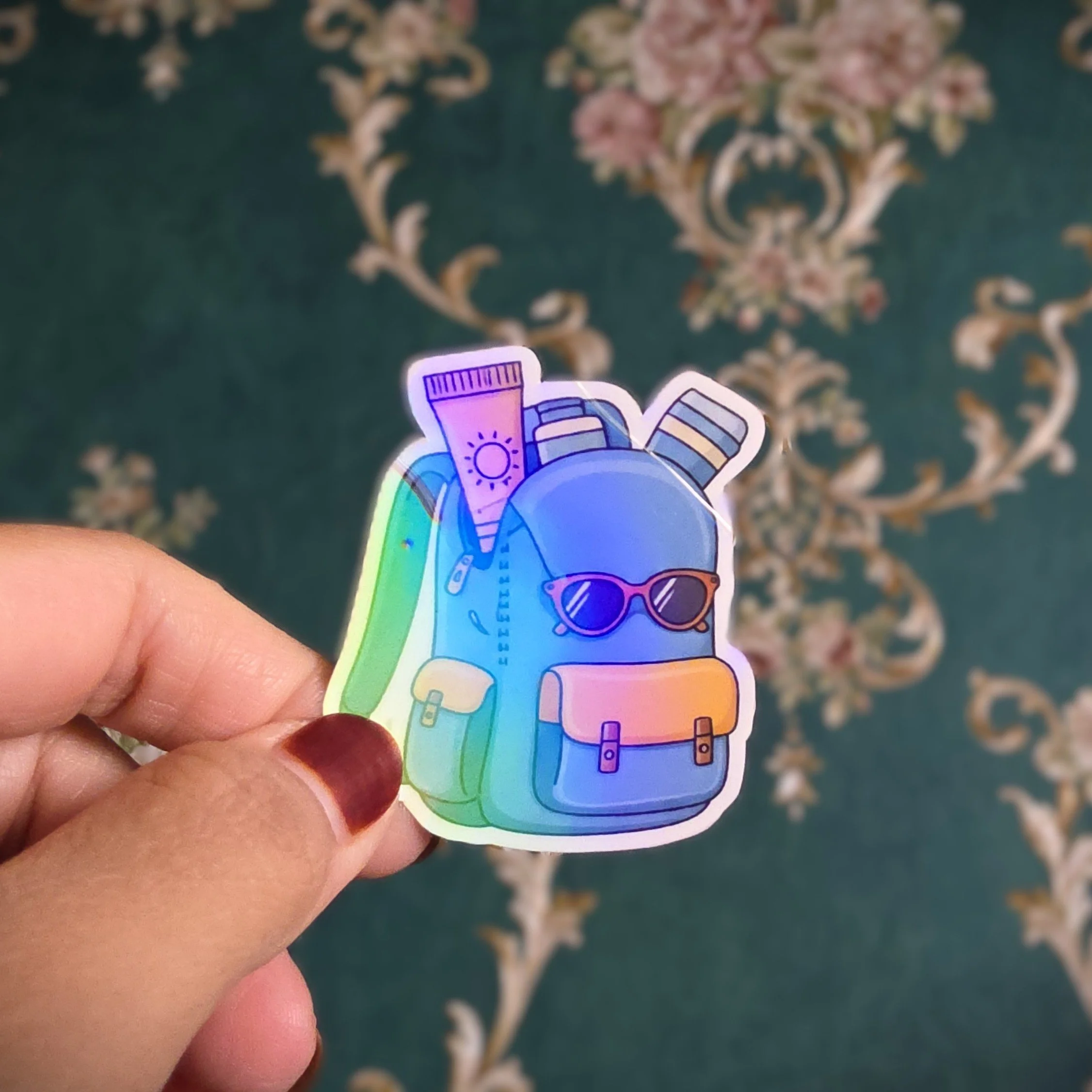 Colorful sticker of a backpack with sunglasses, a sunscreen tube, and a magazine, held in a person’s hand.