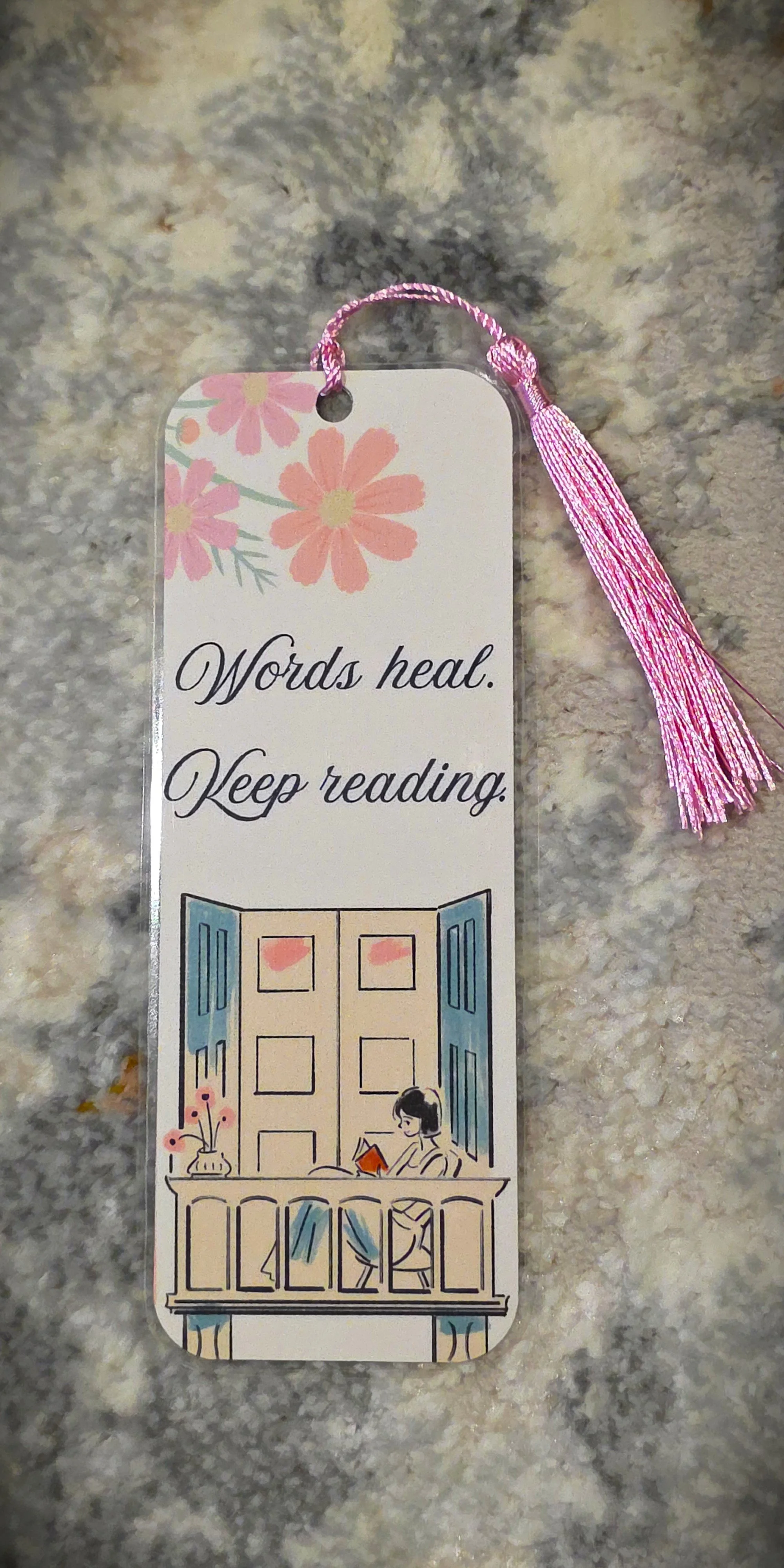A decorative bookmark featuring a floral design at the top, the quote "Words heal. Keep reading." in cursive, and an illustration of a woman reading on a balcony with open blue shutters and a table with a flower vase.