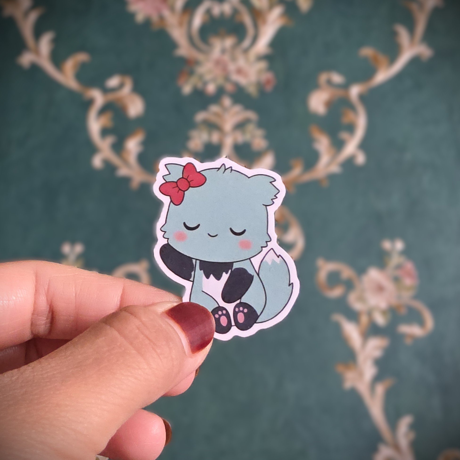A hand holding a sticker of a cute, cartoon-style gray fox with a red bow on its head, sitting with its eyes closed and paws together.