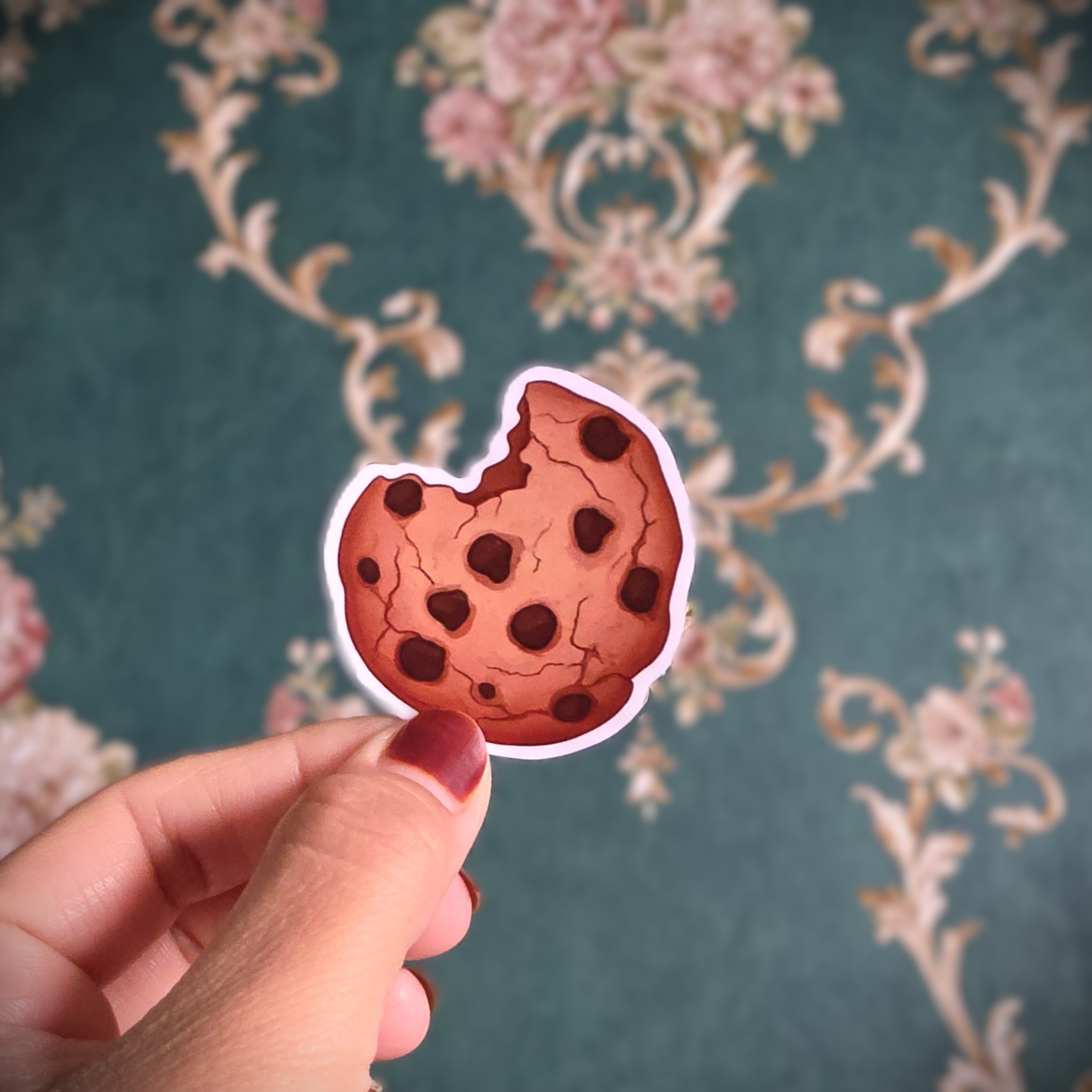 Hand holding a sticker of a chocolate chip cookie with a bite taken out, in front of a decorative floral-patterned carpet.