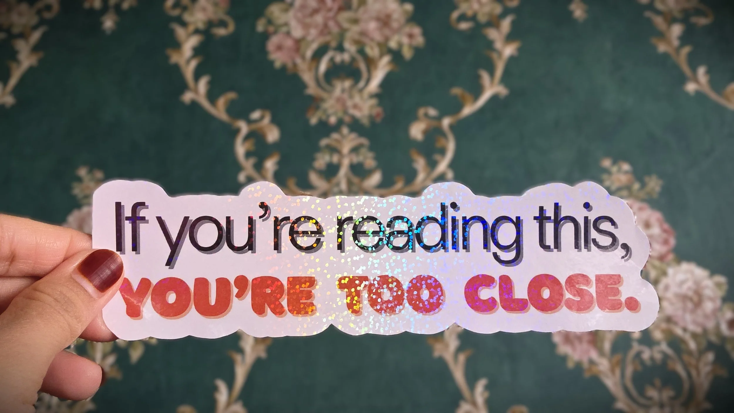 A hand with maroon painted fingernails holding a decorative sticker that says, "If you're reading this, you're too close," with holographic and multicolored text, against a patterned fabric background.