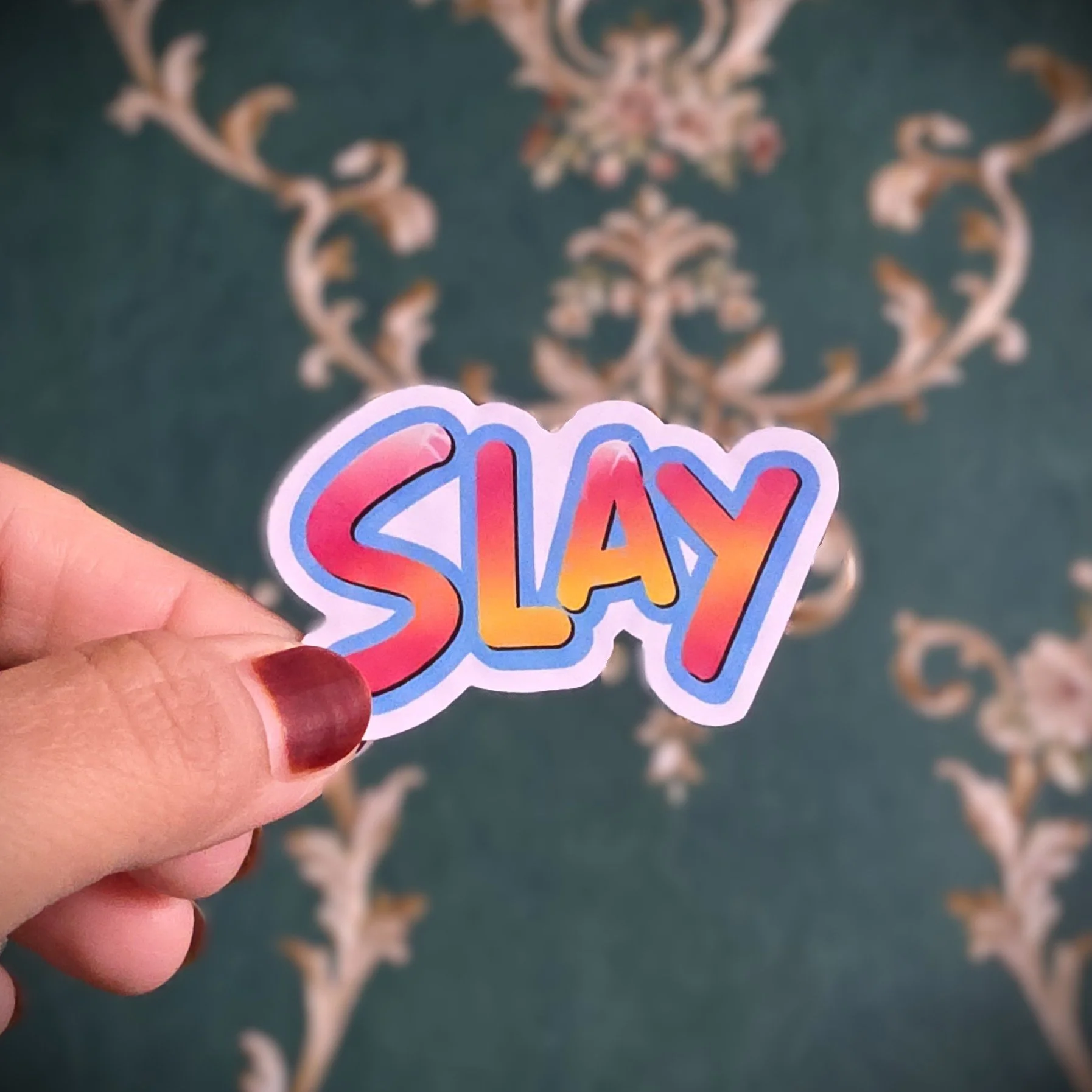 A hand with red-painted nails holding a sticker that says 'SLAY' in colorful, gradient text, with a decorative rug featuring floral and vine patterns in the background.