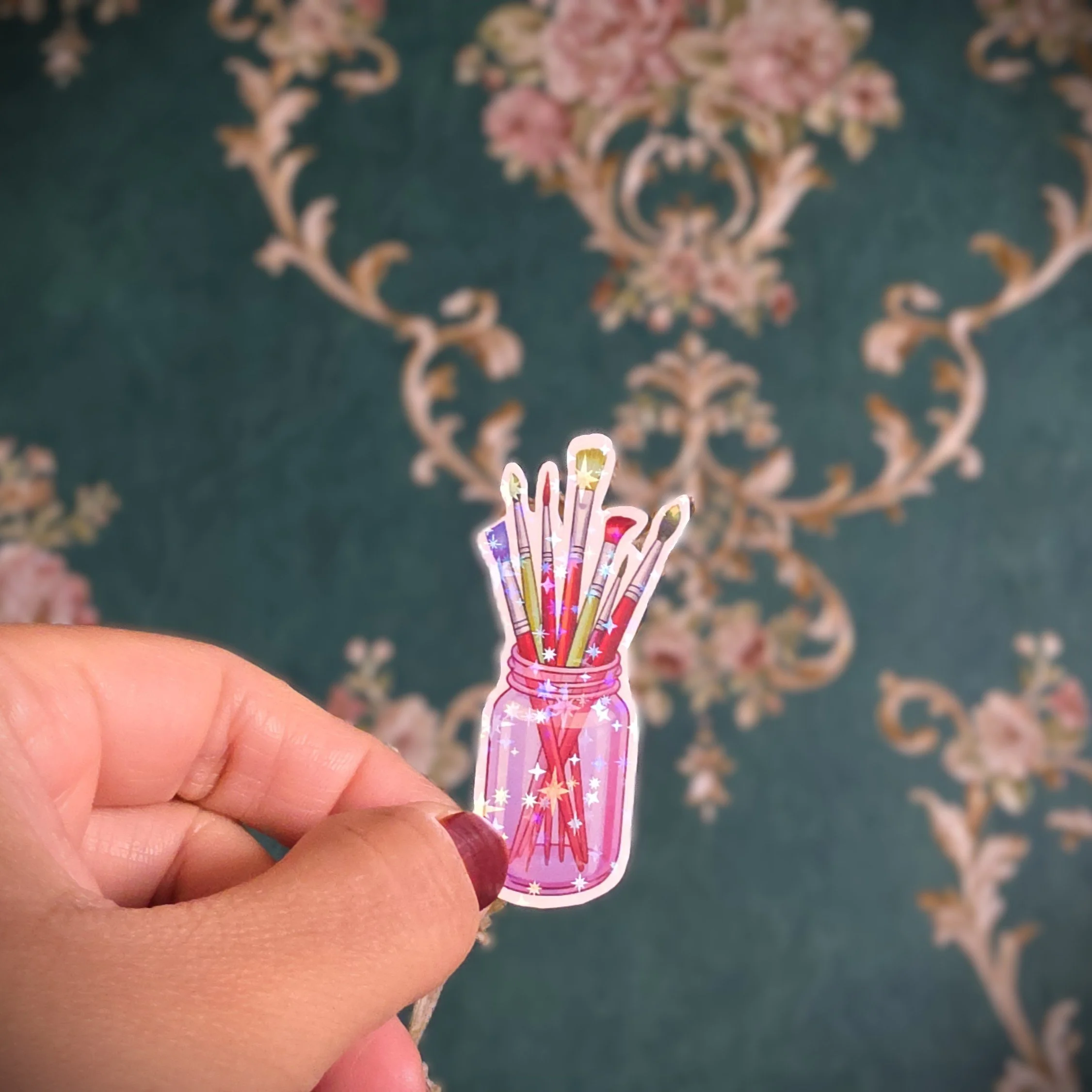 A hand holding a sticker of a pink jar with colorful sparkles, containing five glittery birthday candles, against an ornate, floral-patterned wallpaper background.