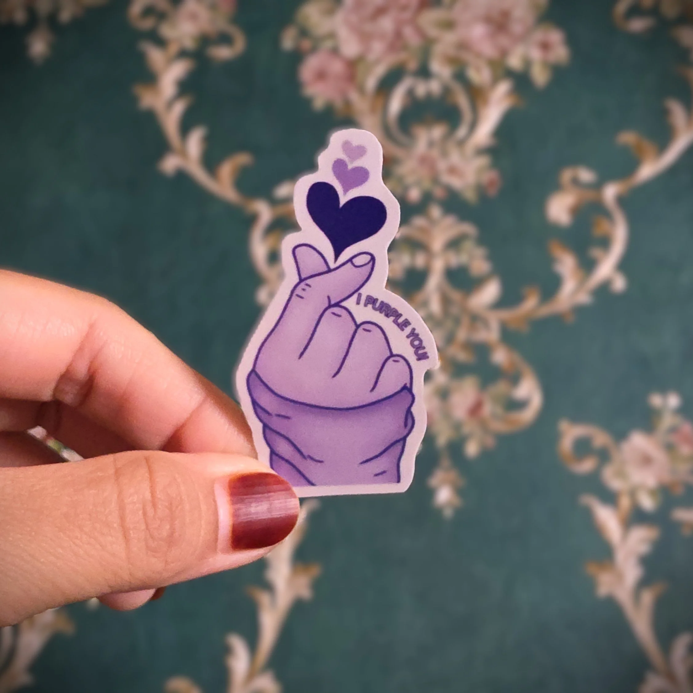 A hand holding a purple sticker with a cartoon-style hand gesture making a finger heart, emitting purple hearts with the words 'I FUMBLE YOU' on it. The background is a green and beige ornate patterned carpet.