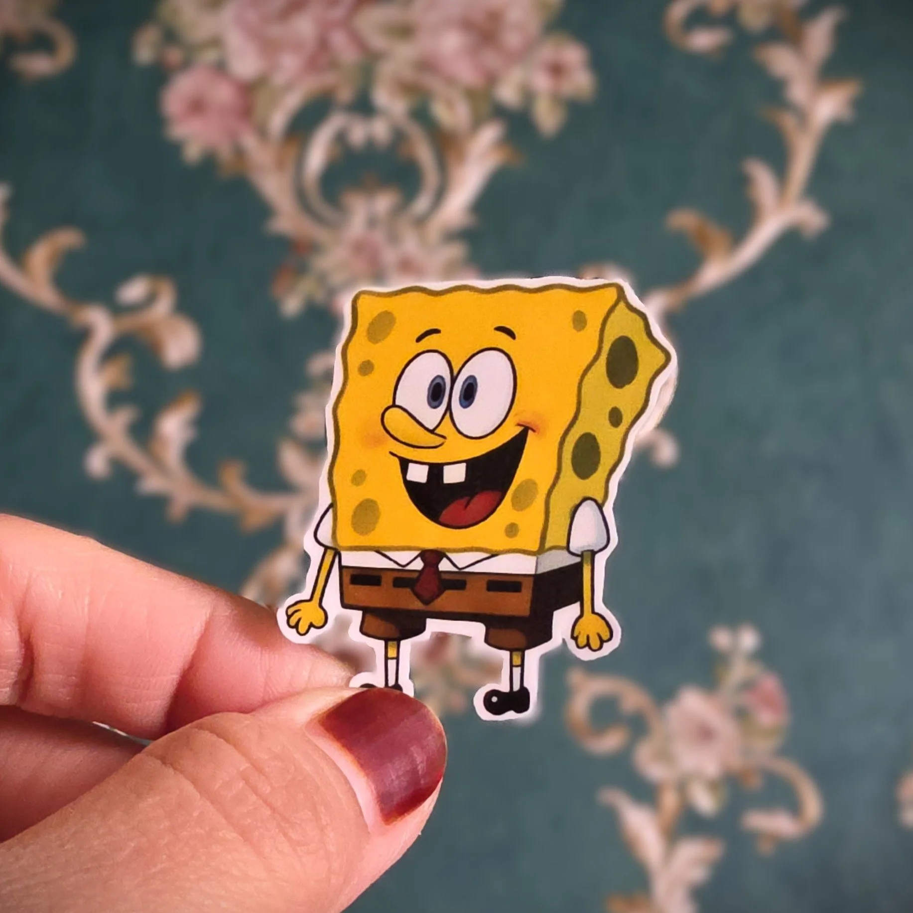 A person holding a SpongeBob SquarePants sticker with a floral-patterned carpet in the background.