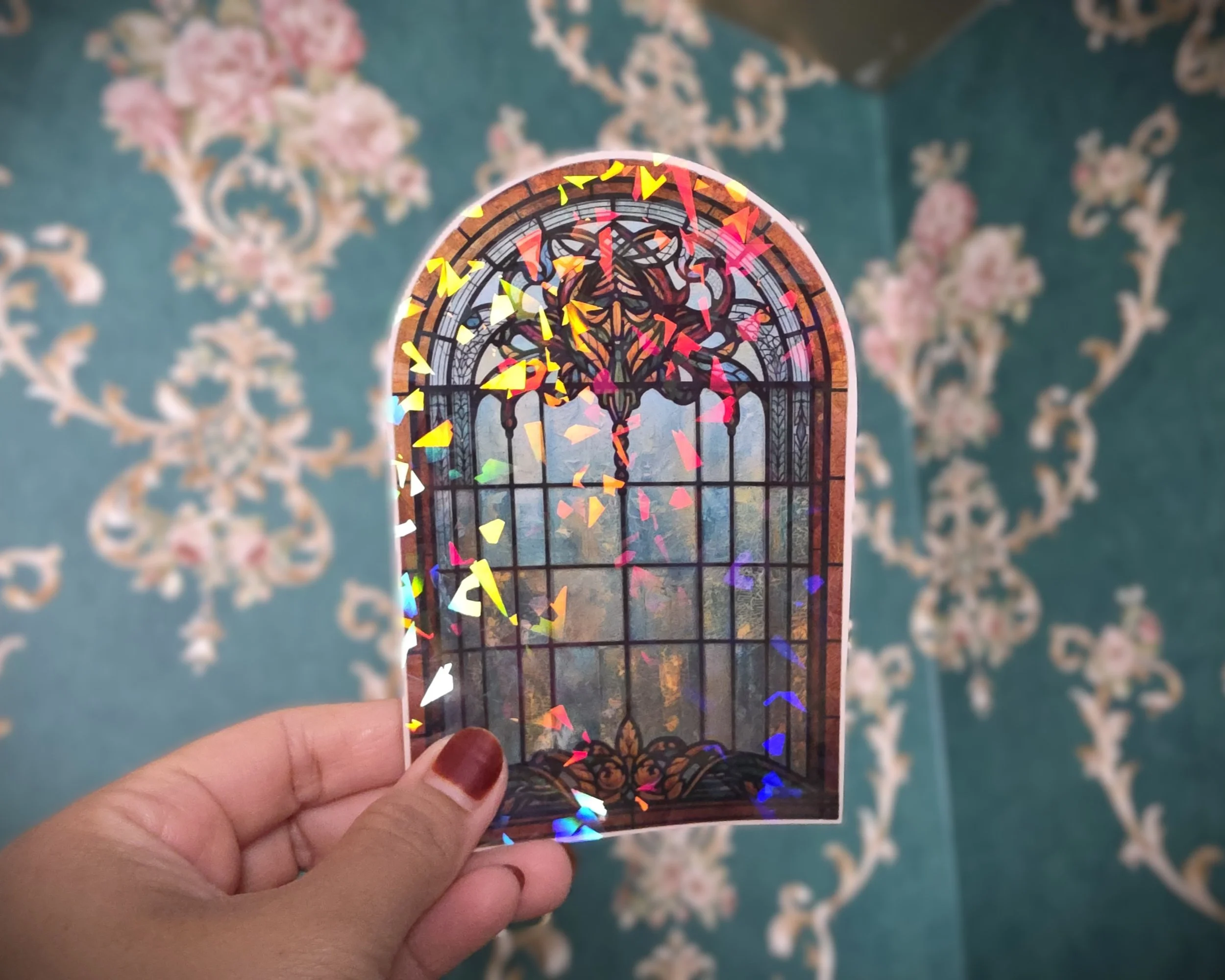 A hand holding a small, arched stained glass window with colorful, reflective confetti outside it. The background is a teal wall with pink and white floral patterns.