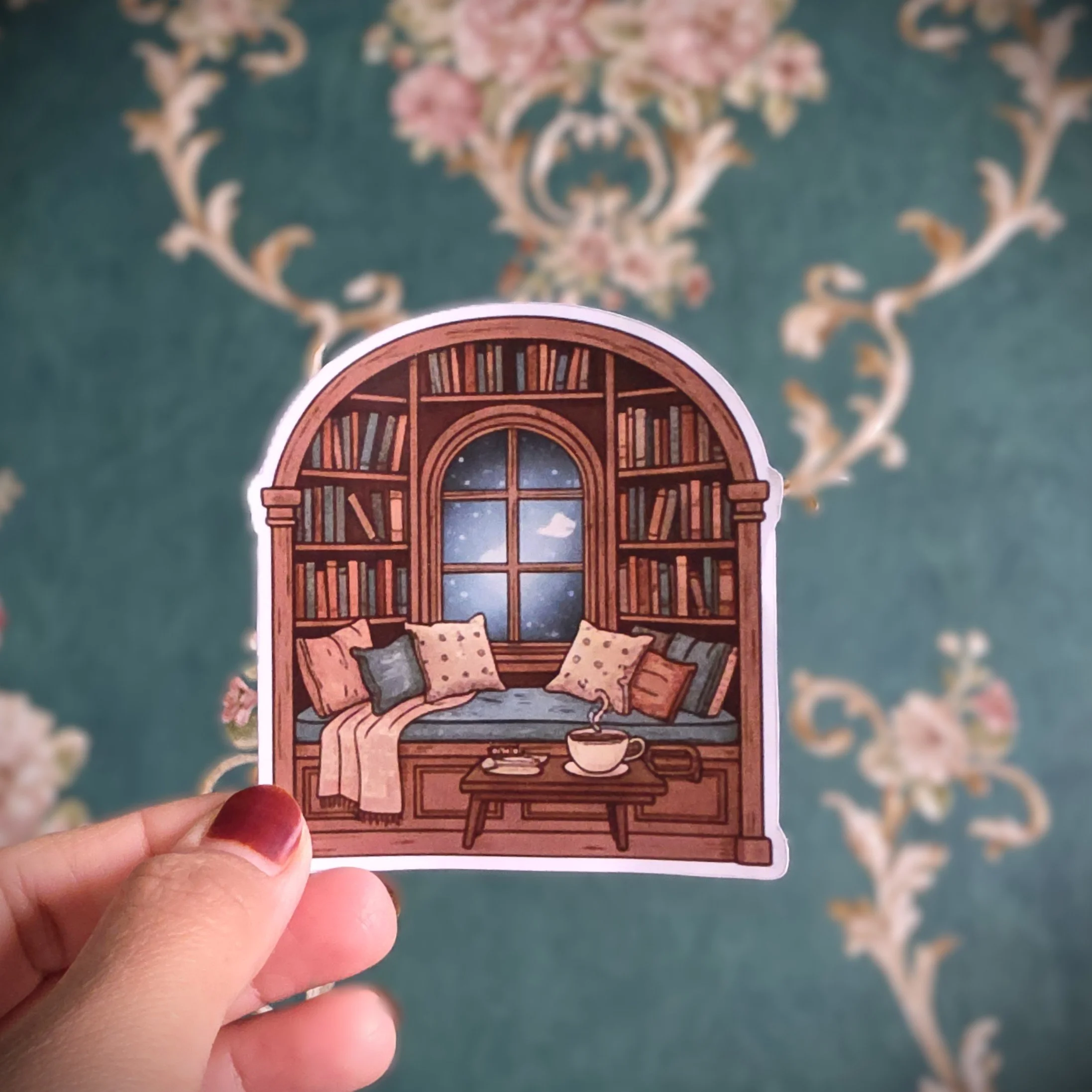 Hand holding a sticker of a cozy library nook with a window showing a starry night sky, bookshelves, a window seat with pillows, a steaming cup, and a small table.