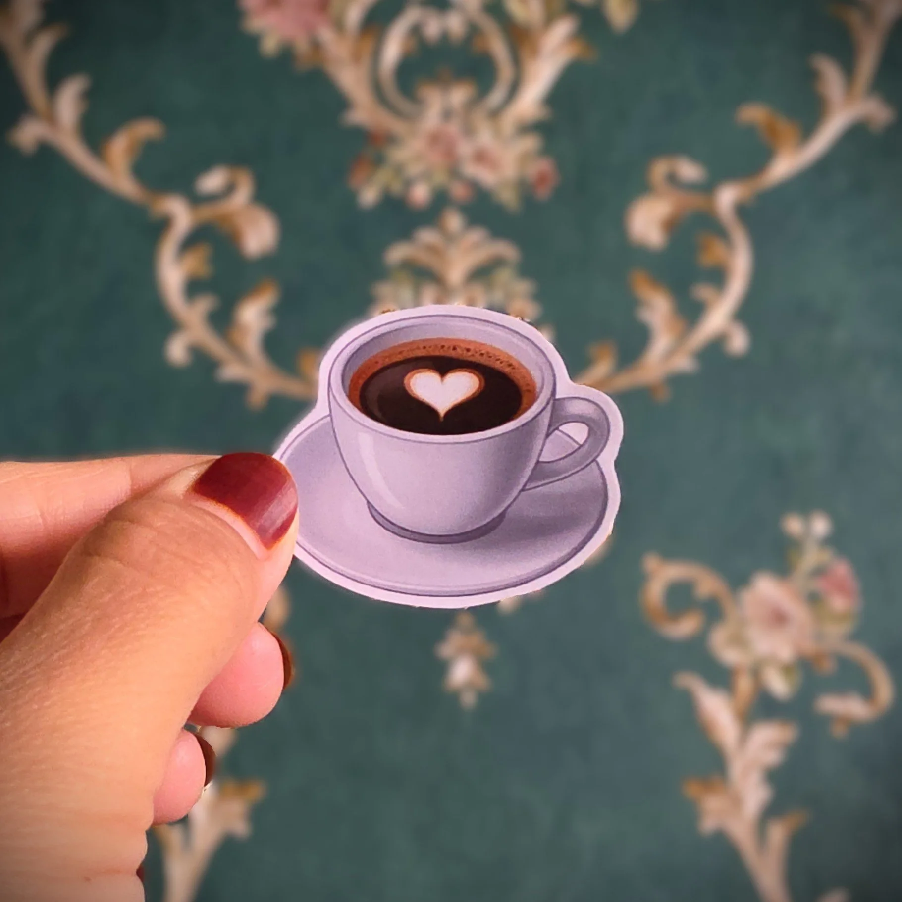 Photo of a small sticker of a white coffee cup with a saucer, filled with black coffee with a heart-shaped foam on top, held between fingers with a painted nail, against a patterned green and gold wallpaper background.