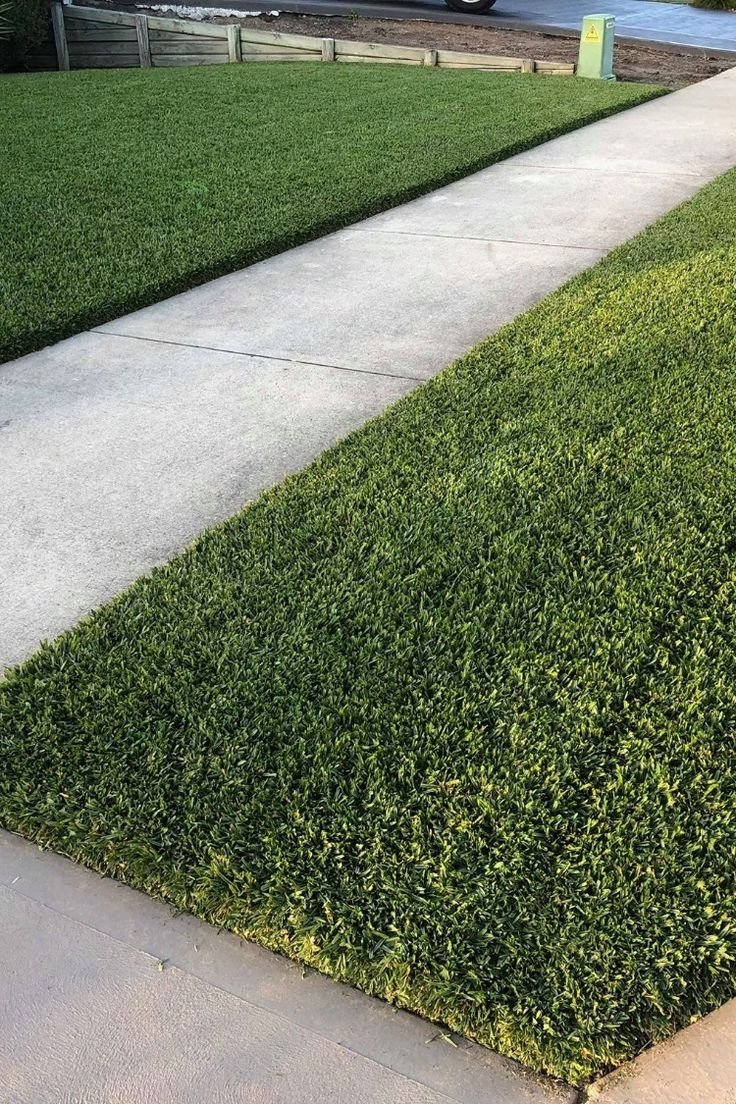 A sidewalk running between two areas of neatly trimmed green grass, with a wooden fence and utility box in the background.