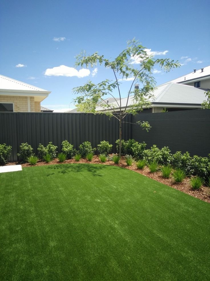 A backyard with lush green grass, a small tree, green bushes along a dark gray fence, and a clear blue sky with a few clouds.