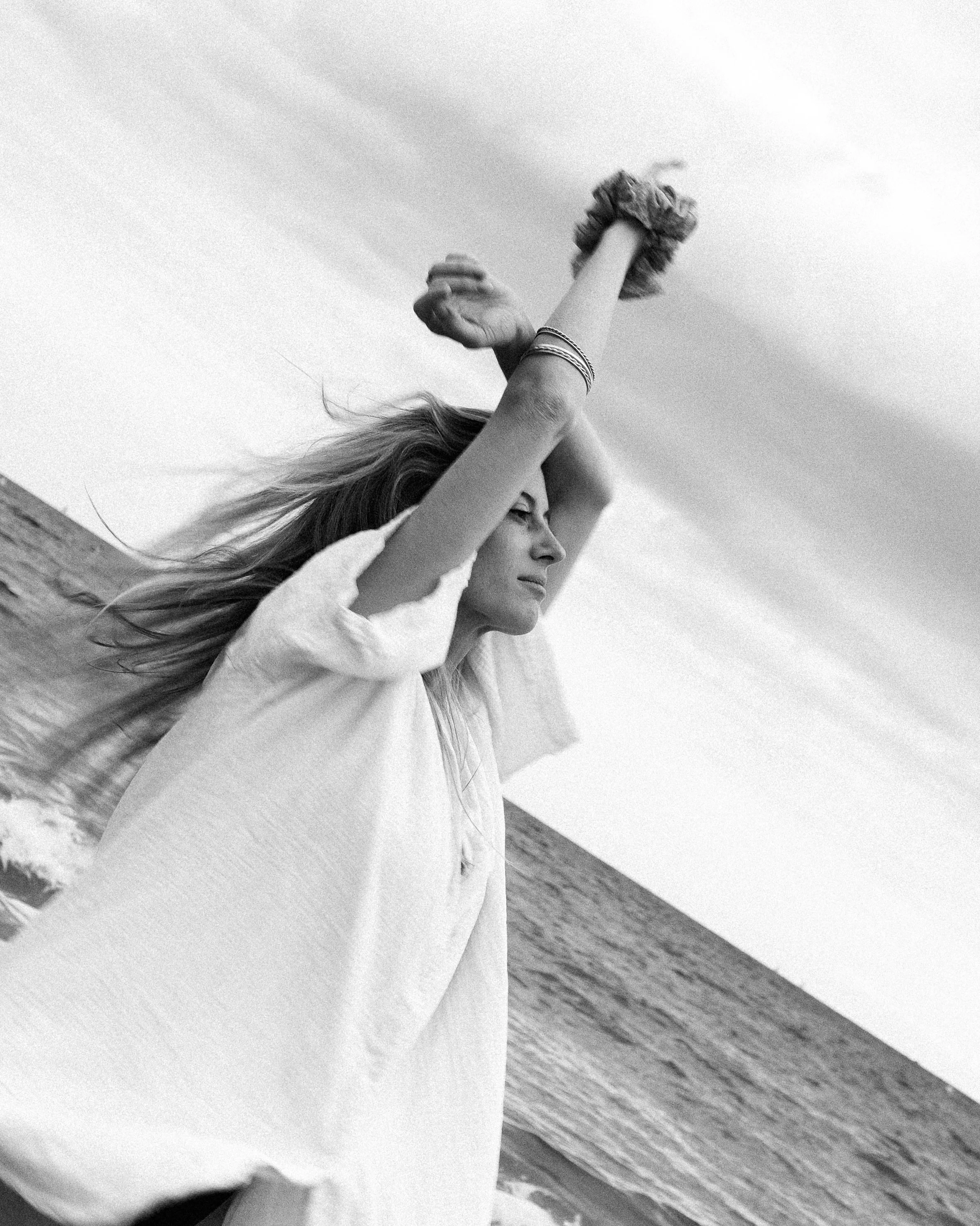 Une femme blonde porte une robe légère et des bracelets, elle se tient sur la plage face à la mer, levant les bras en l'air dans un mouvement de liberté.