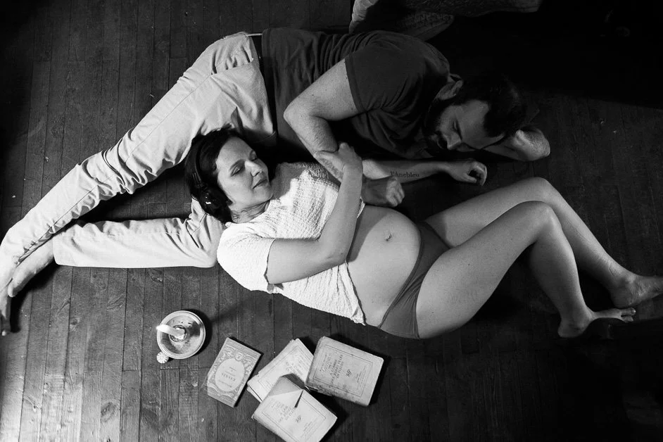 Une photographie en noir et blanc d'un couple allongé sur un sol en bois. La femme porte un T-shirt léger et des sous-vêtements, elle est allongée sur le côté. L'homme est allongé derrière elle, avec la tête tournée vers le haut, portant un vêtement sombre. Il y a un livre ouvert et deux autres livres empilés, ainsi qu'une bougie dans un contenant autour d'eux.