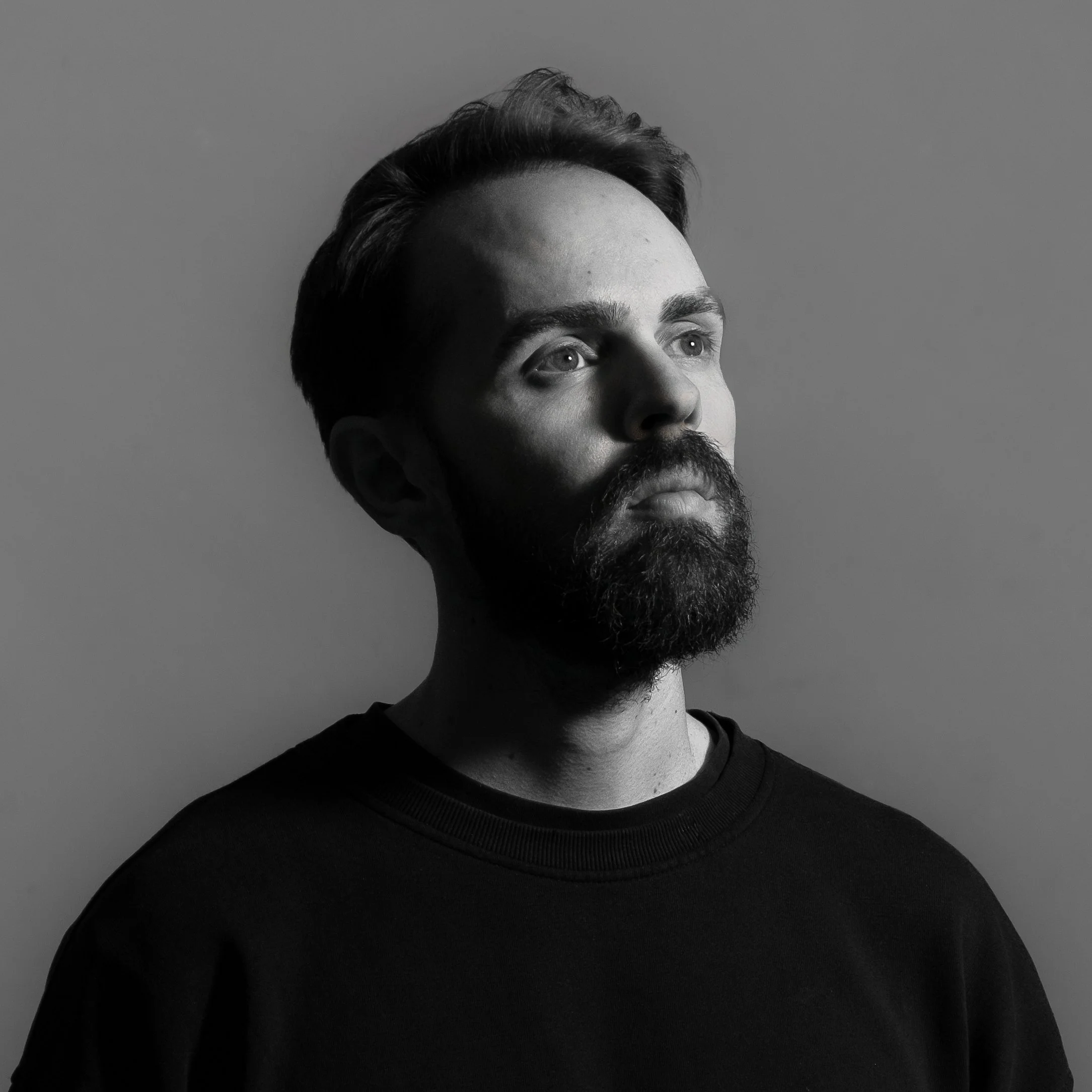 Un portrait en noir et blanc d'un homme avec une barbe, regardant vers le haut, fond gris clair, éclairage doux