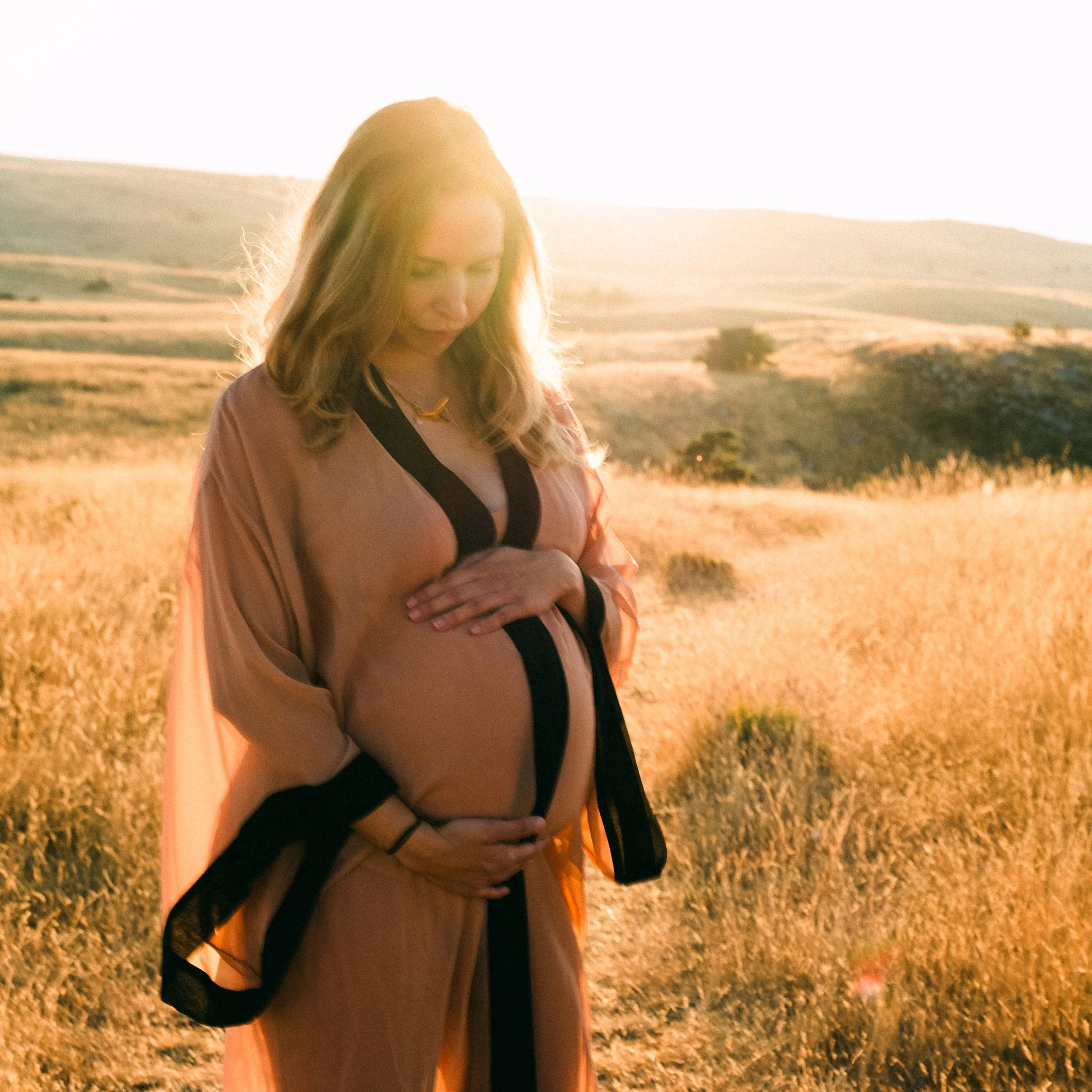 Femme enceinte dans un champ doré au coucher du soleil, portant une robe beige avec des détails noirs.