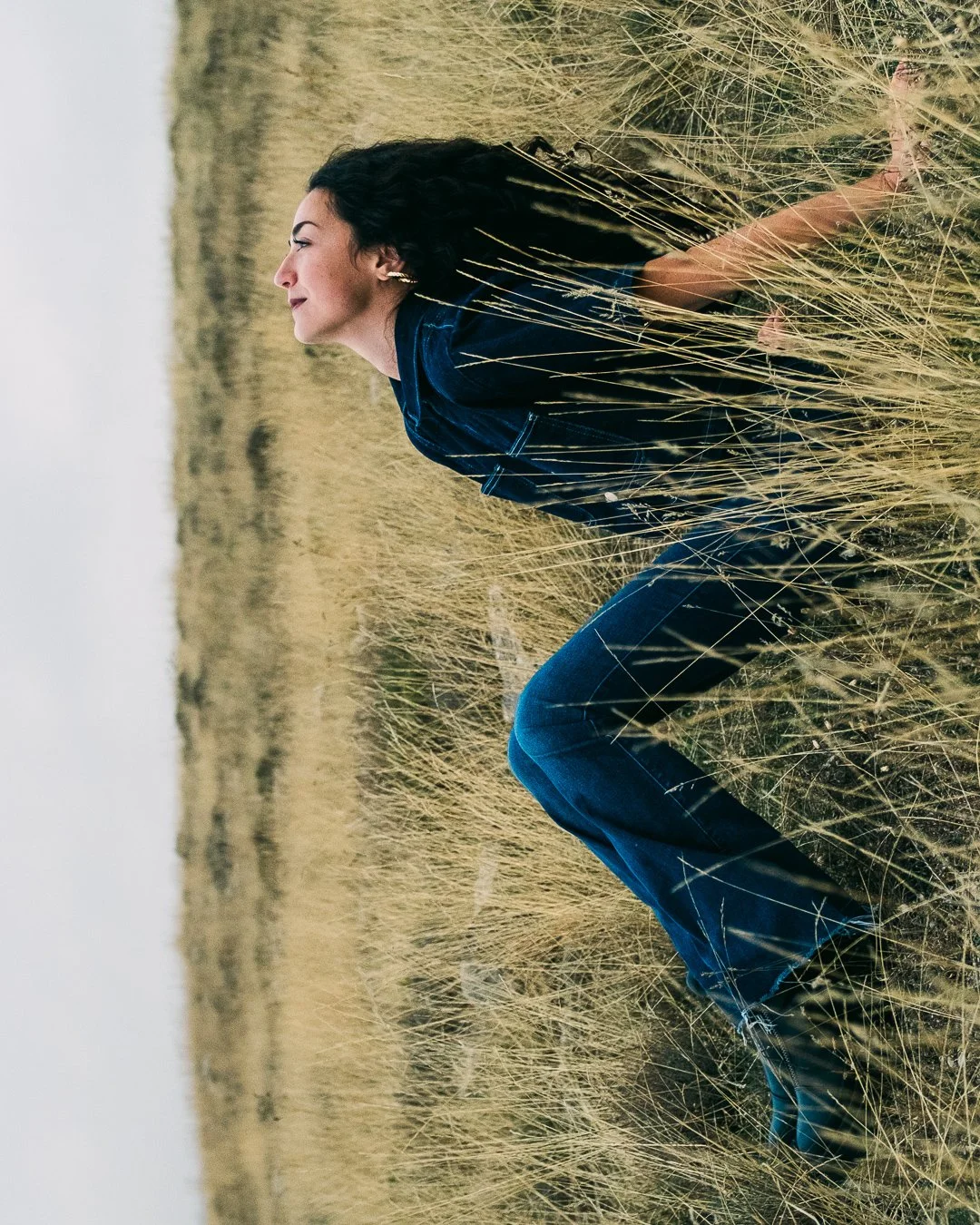 Femme assise dans un champ de herbes sèches, regardant vers la gauche, portant un jean et un haut bleu, avec un ciel nuageux en arrière-plan.