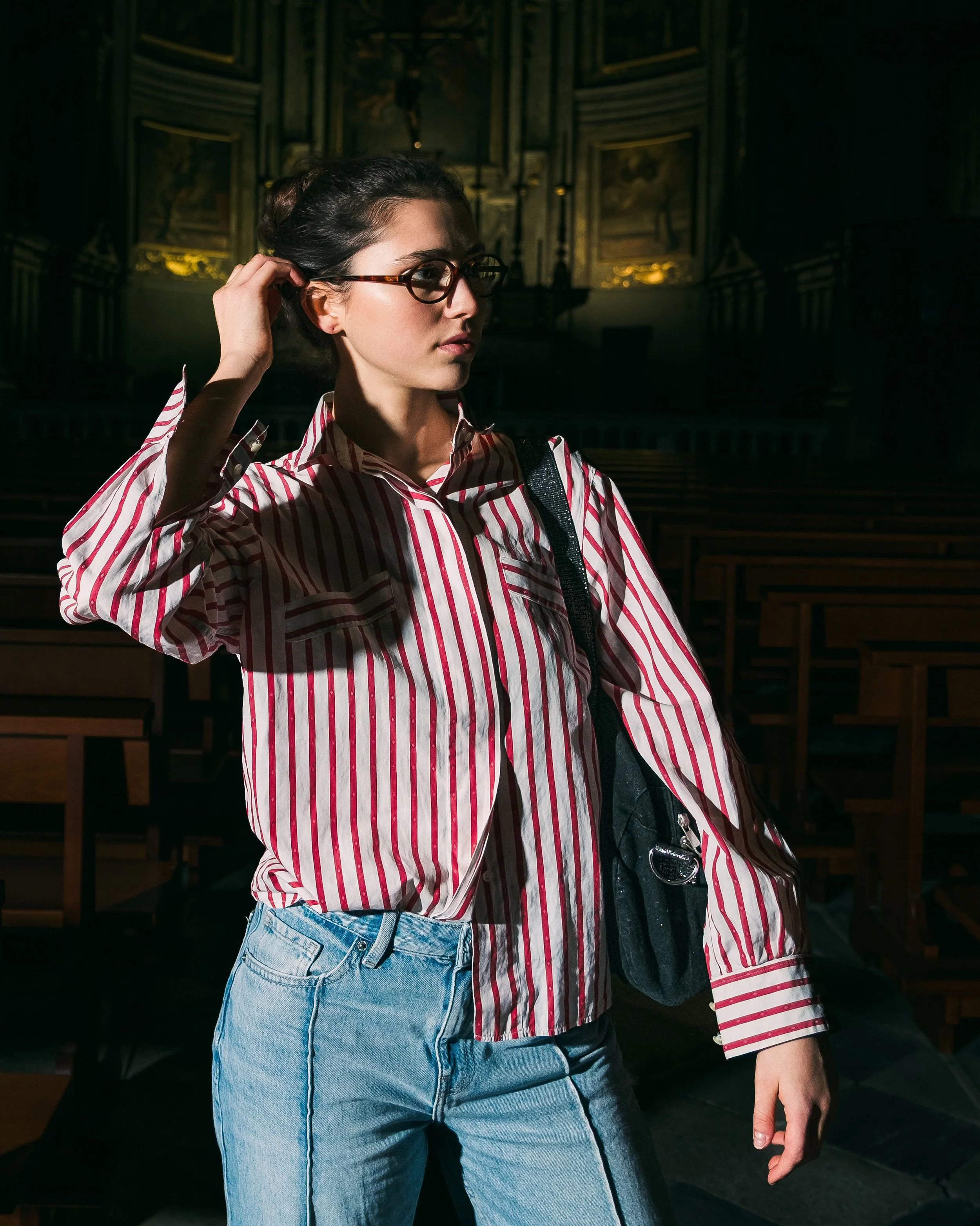 Jeune femme portant des lunettes, une chemise à rayures rouges et blanches, et un sac à l'épaule, debout dans une salle sombre avec des bancs en bois.