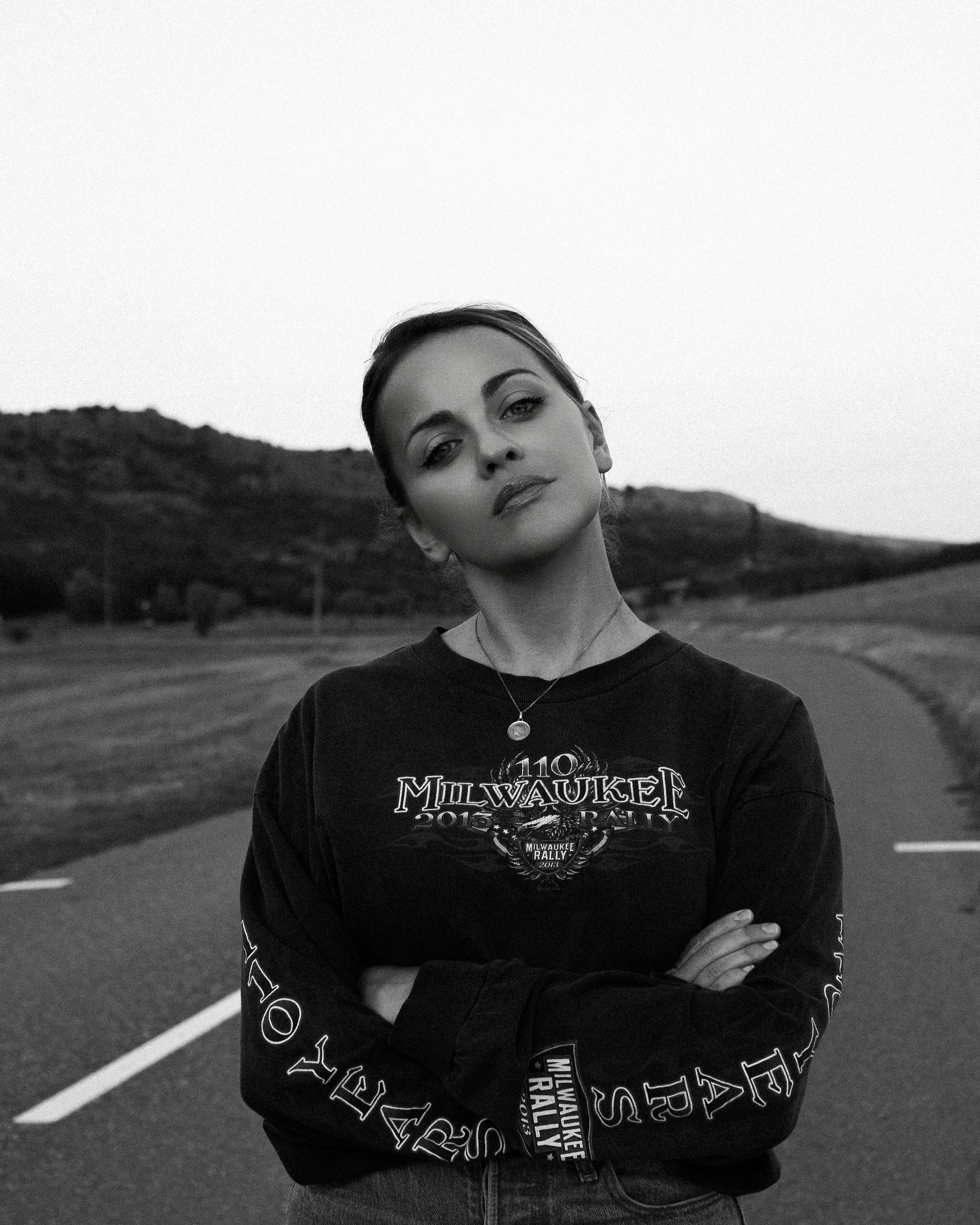 Portrait en noir et blanc d'une femme avec une expression sérieuse, bras croisés, portant un sweat-shirt avec le texte 'Milwaukee 2015 Rally', sur une route rurale avec des collines en arrière-plan.