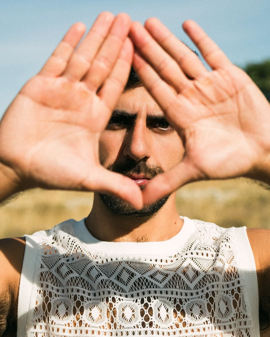 Un homme avec une barbe, portant un haut en dentelle blanc, regarde à travers un cadre formé par ses mains, en extérieur lors d'une journée ensoleillée.
