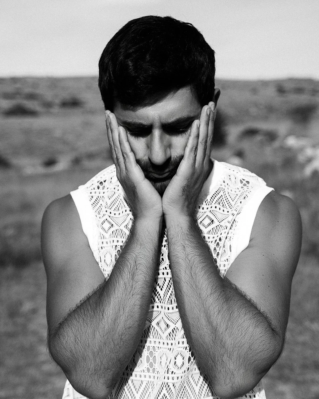 Un homme en noir et blanc, avec les mains sur le visage, regarde vers le bas, dans un décor naturel.