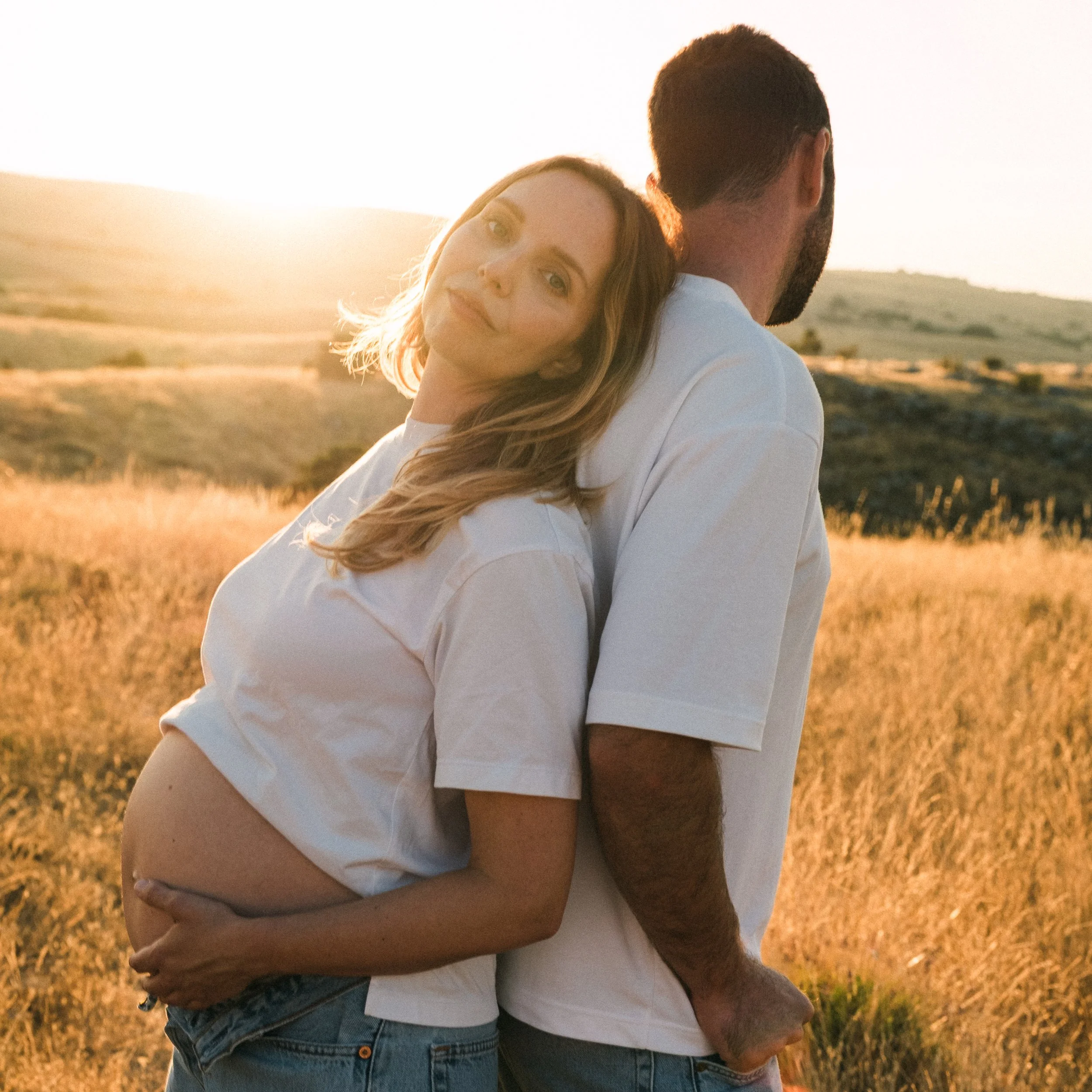 Une femme enceinte et un homme se tiennent dans un champ avec un coucher de soleil en arrière-plan, la femme repose sa tête sur l'épaule de l'homme.