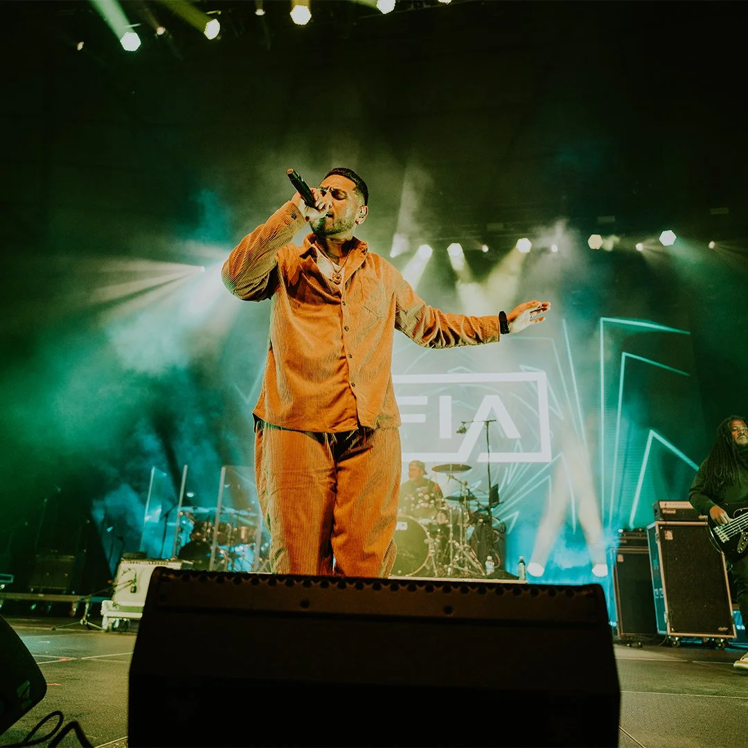 Male singer in tan outfit performing on stage with a band behind him, colorful lighting, and a digital display in the background.