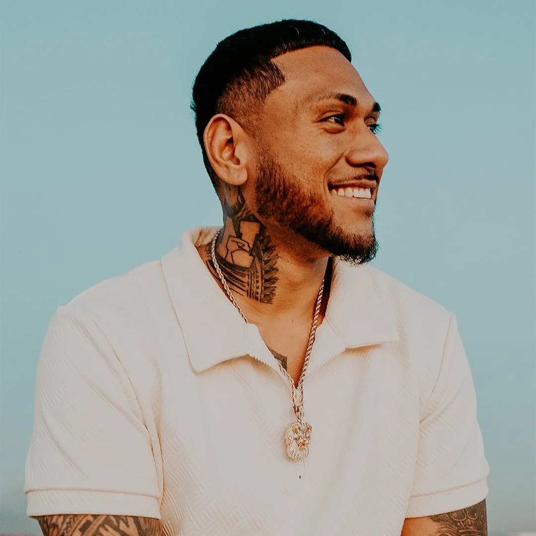 A smiling man with tattoos and a beard, wearing a white shirt and gold jewelry, against a clear blue sky.