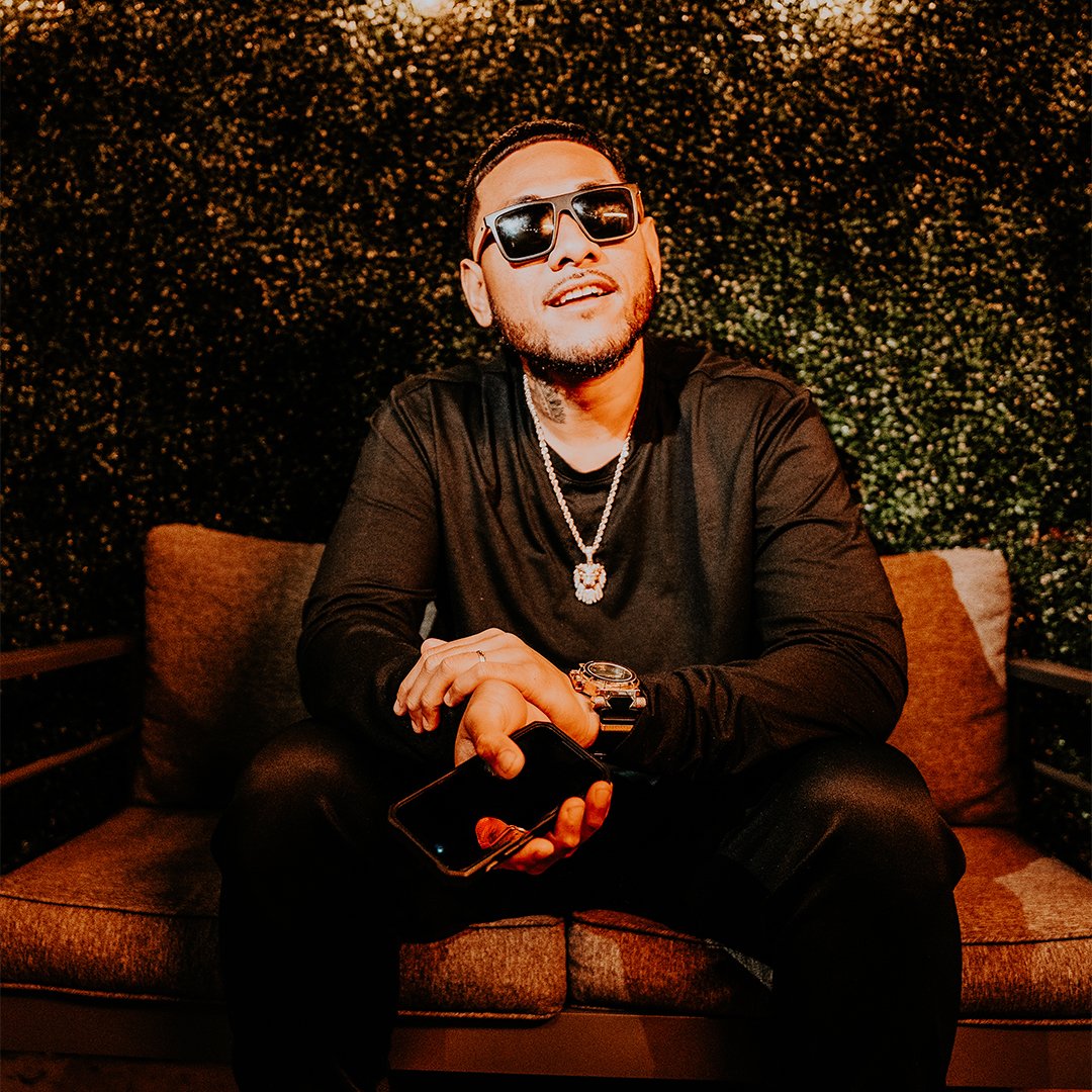 A man sitting on a brown outdoor chair at night, wearing black sunglasses, a black long-sleeve shirt, a silver chain necklace, and a watch, holding a smartphone, with a green hedge background.