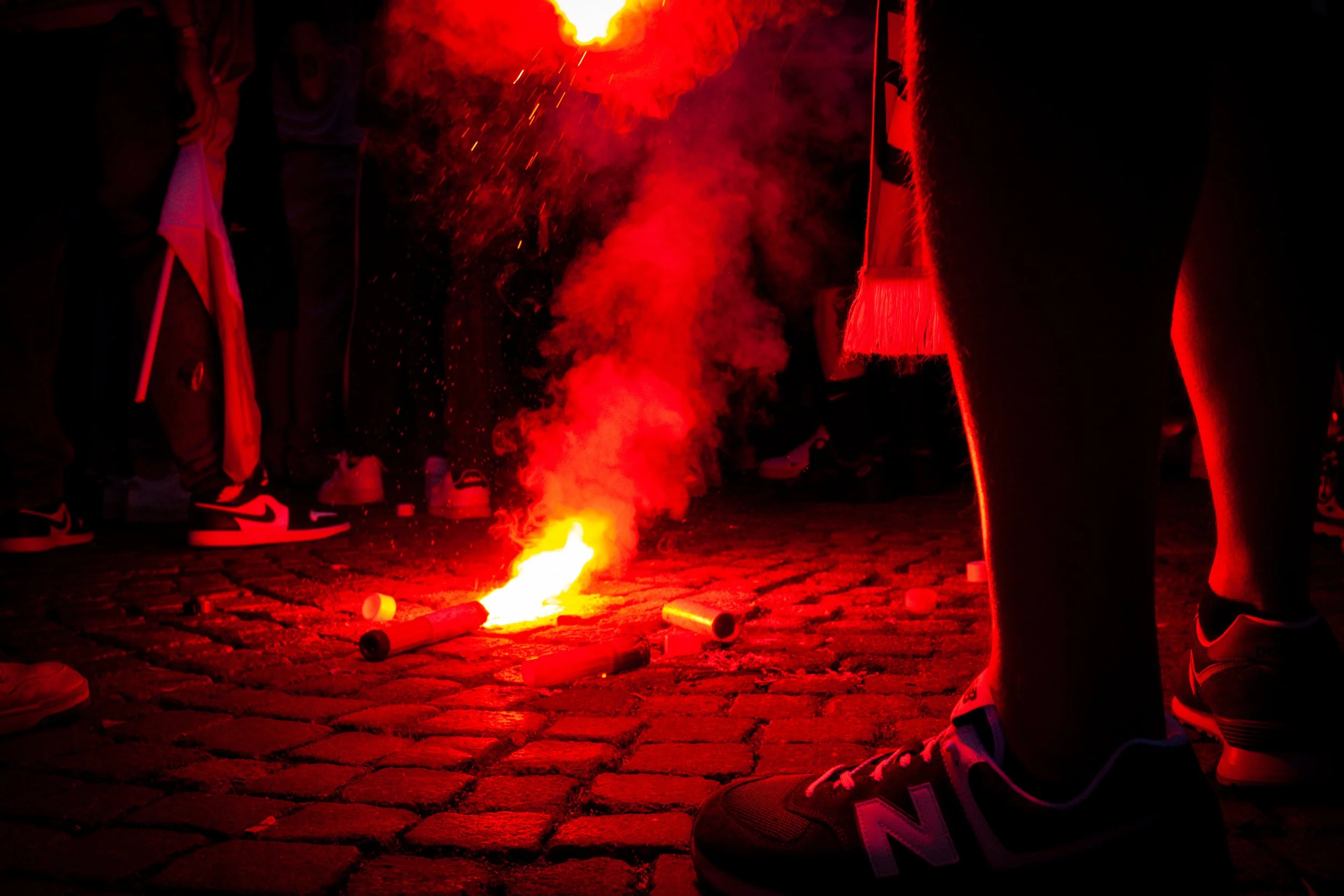 Persone con sneakers che partecipano a una manifestazione, con un fuoco artificiale acceso sul pavimento di pietra, accompagnato da fumo rosso.